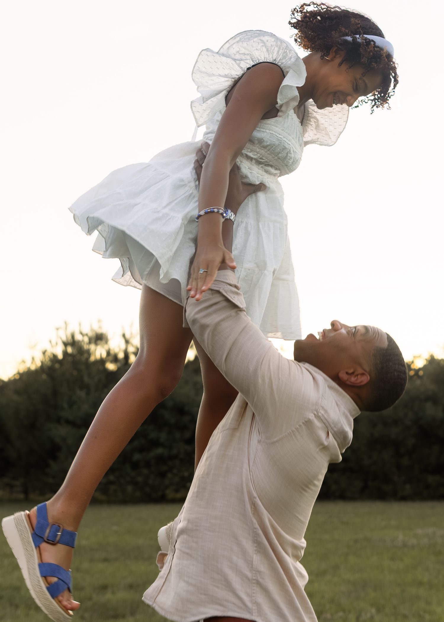 A parent joyfully lifts a young girl in a white dress outdoors on a green lawn during golden hour.