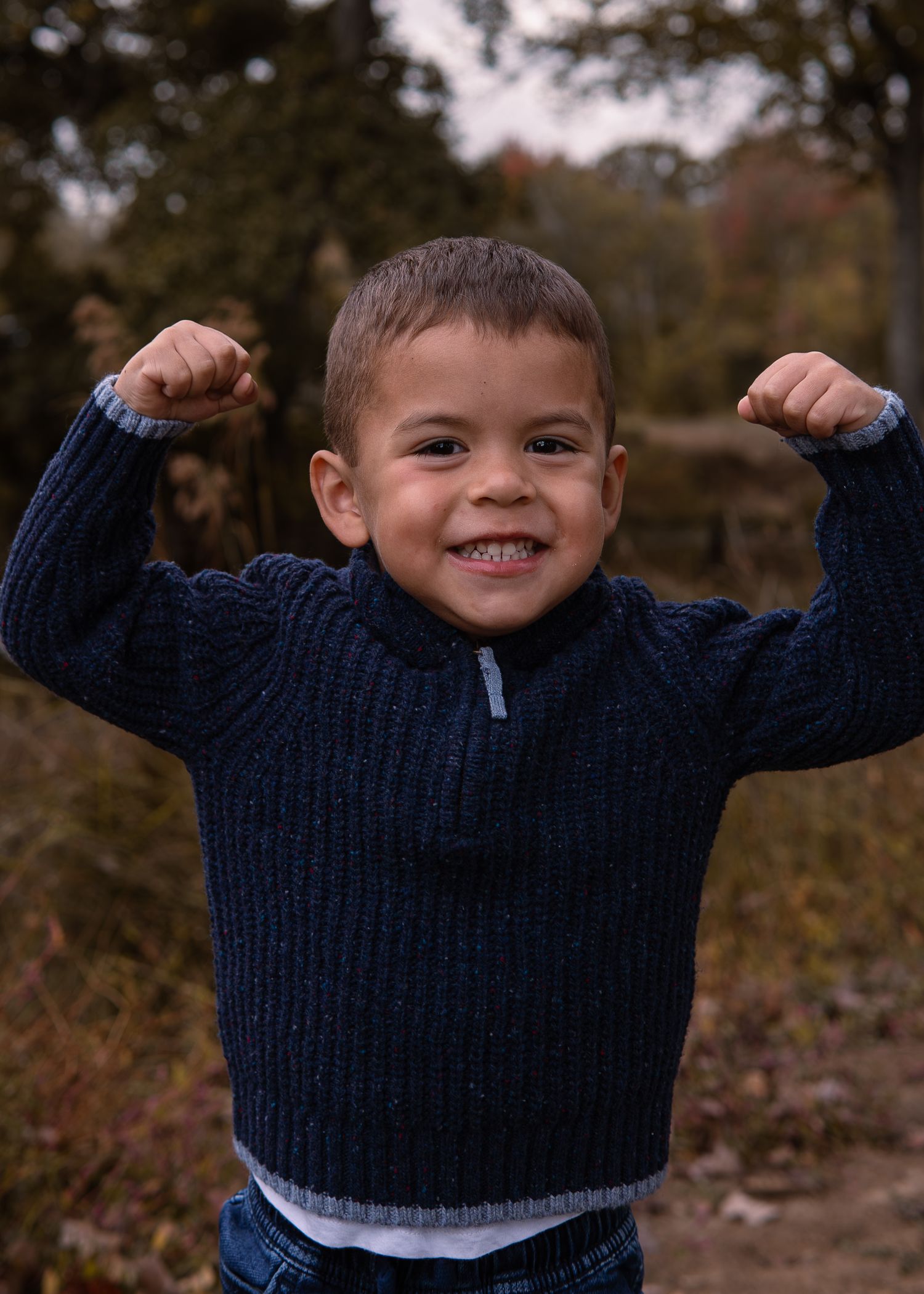 Young boy in a navy knit sweater flexing both arms and smiling proudly in an autumn outdoor setting.
