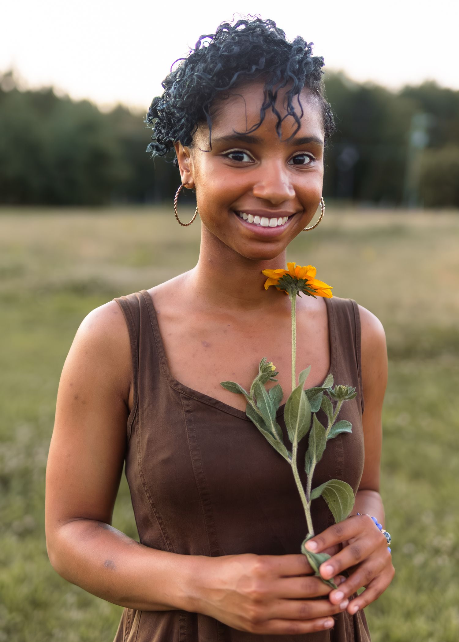 Smiling young woman holding sunflower in brown dress standing in open field at dusk.