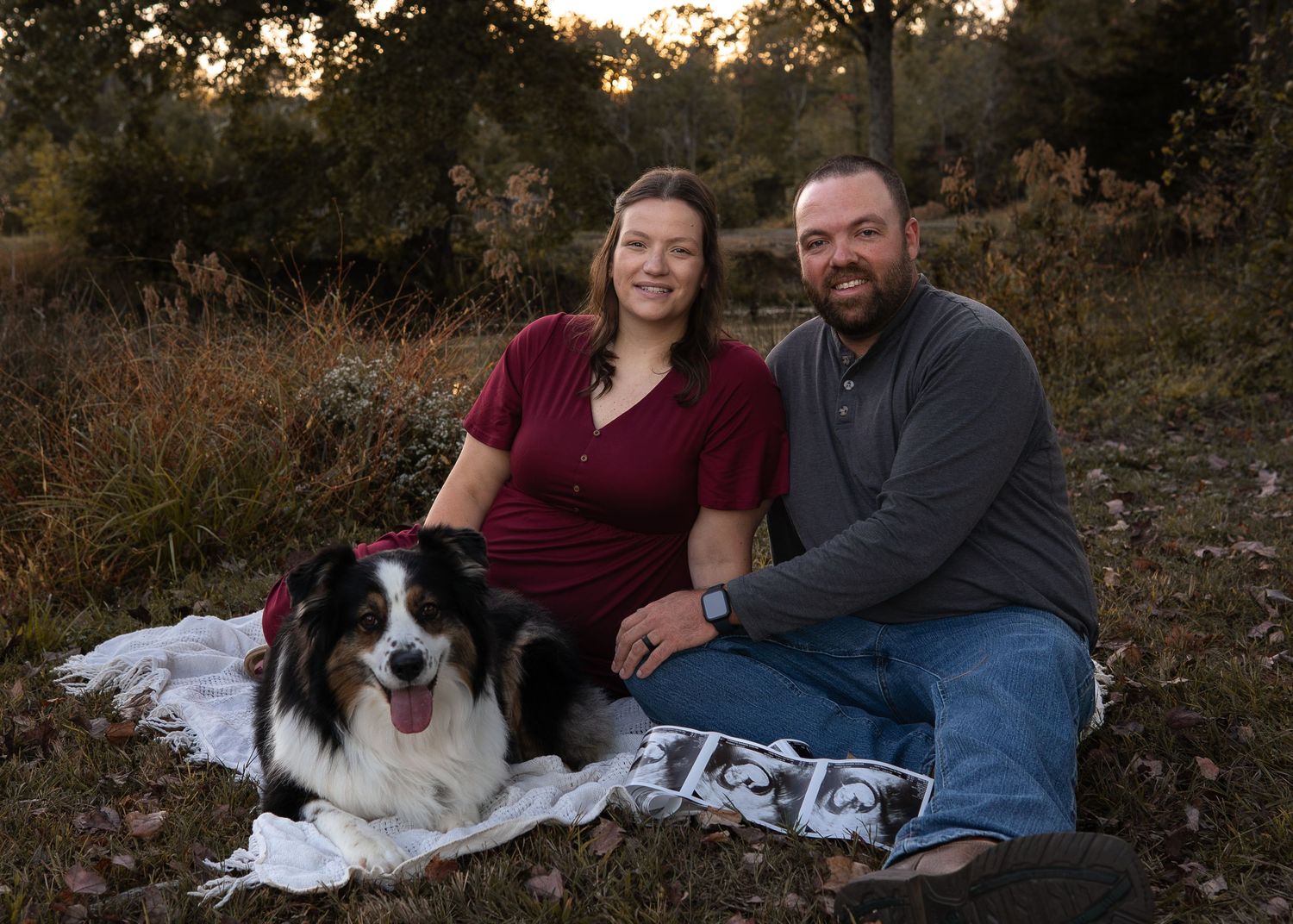 Couple sitting outdoors with their Australian Shepherd dog on a blanket with ultrasound photos at golden hour.