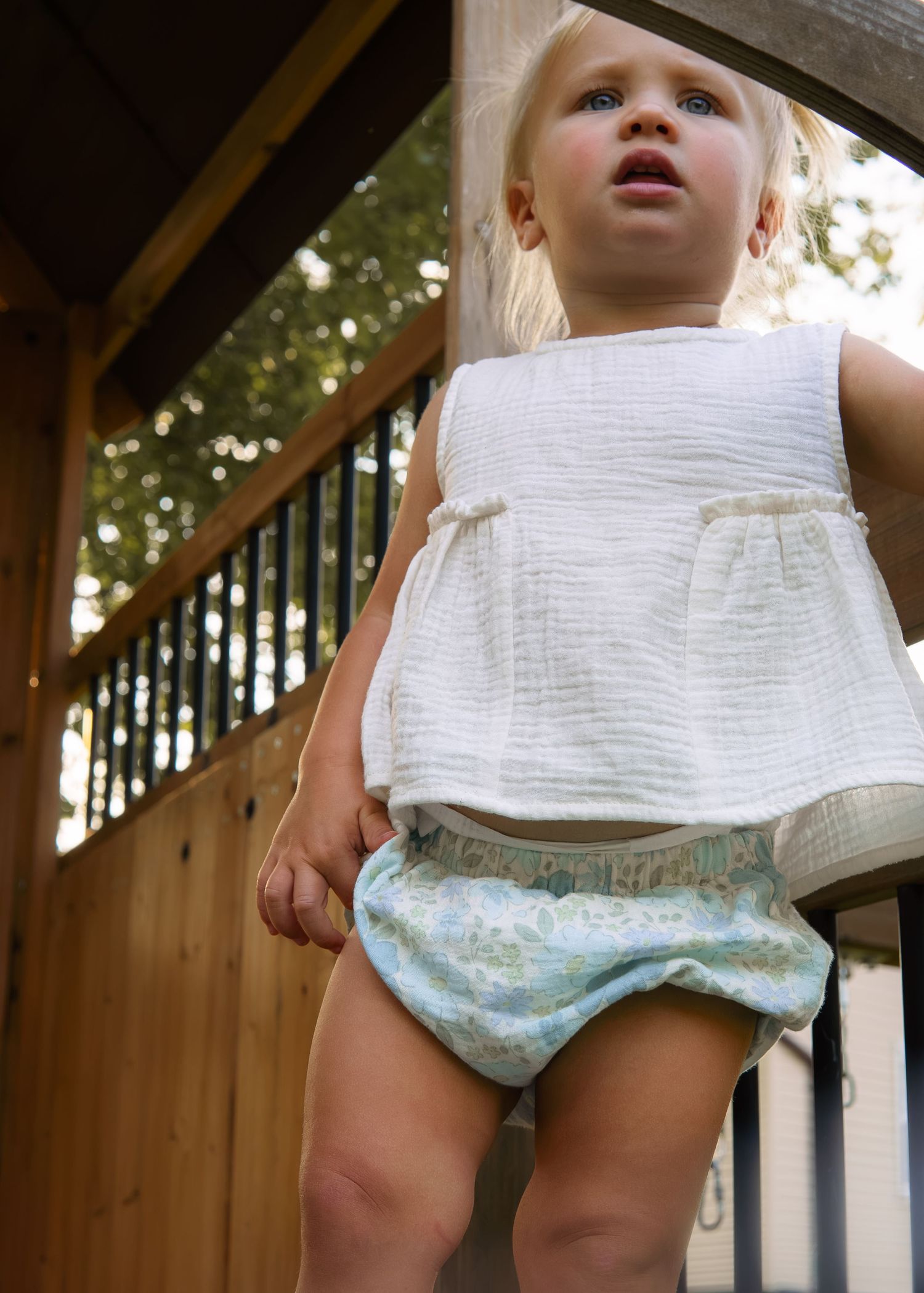 Toddler girl in white top and floral bloomers standing on wooden play structure.