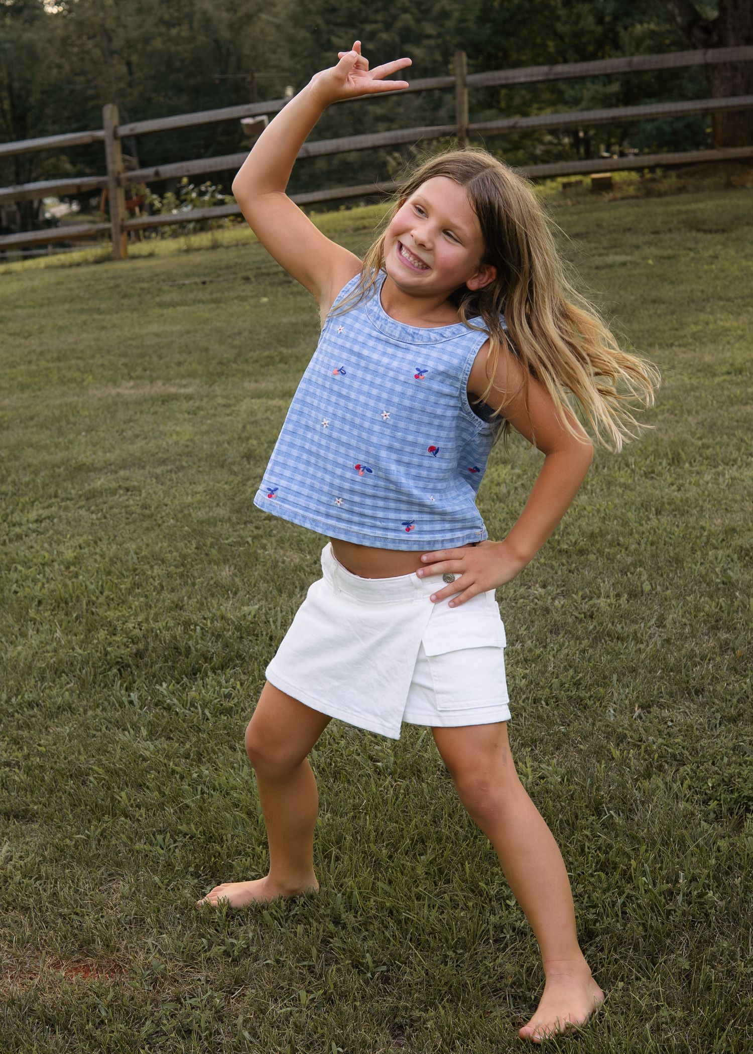 Young girl dancing barefoot in a blue gingham top and white skirt outdoors.