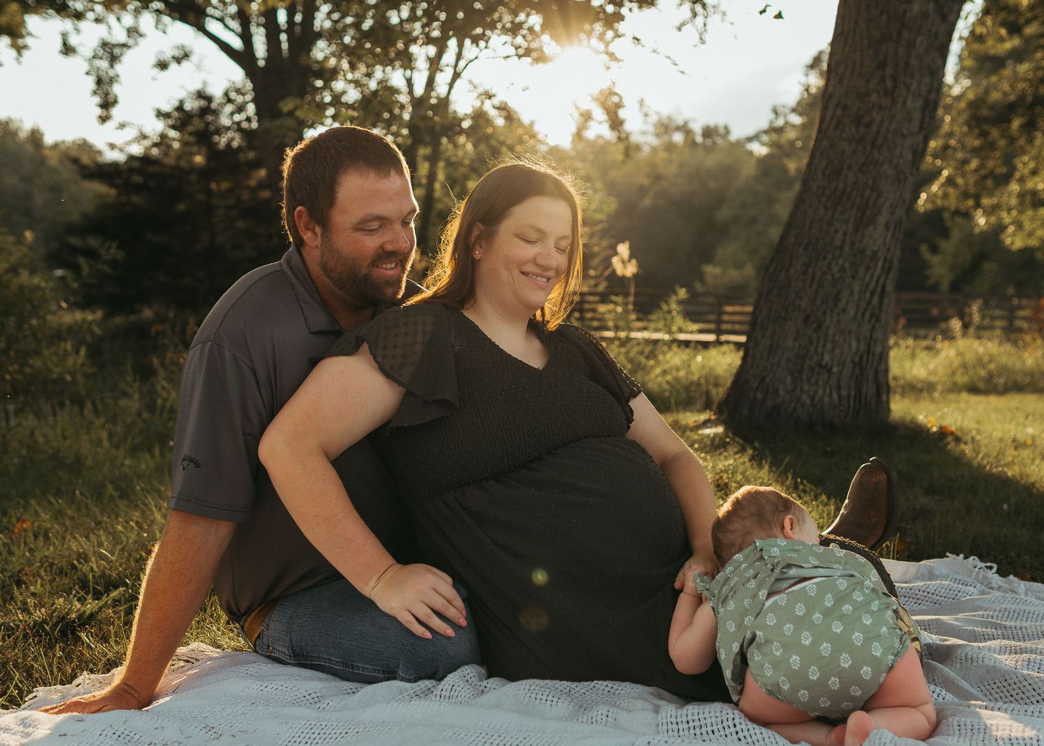 Pregnant couple sitting on blanket outdoors in golden sunlight with toddler leaning on mother's belly.