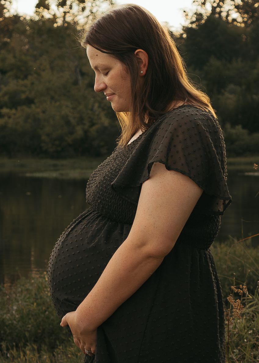 Pregnant woman in black dress standing by a lake at golden hour, looking down at her baby bump.