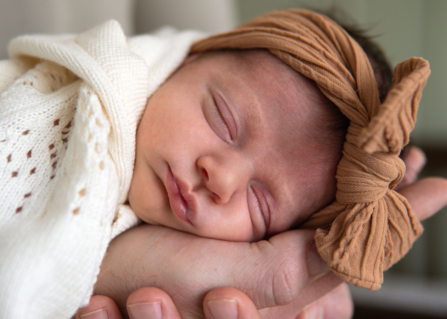 Sleeping newborn baby wrapped in white knit swaddle wearing a caramel bow headband, cradled in adult hands.