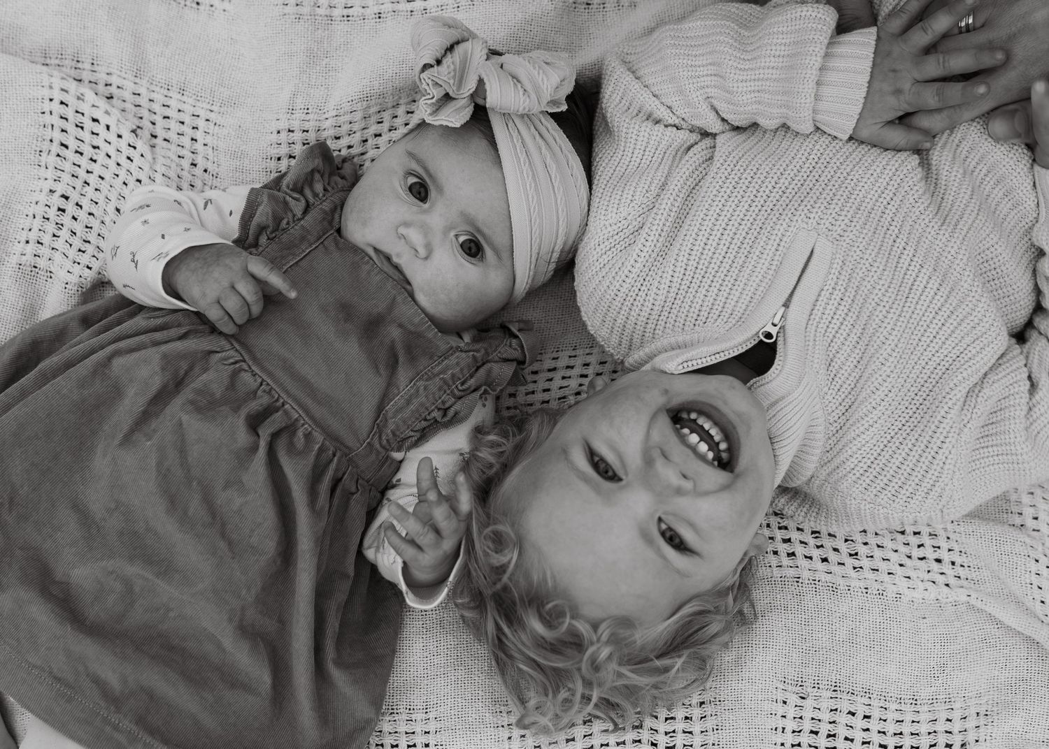 Black and white photo of a baby and toddler lying together on a white blanket.