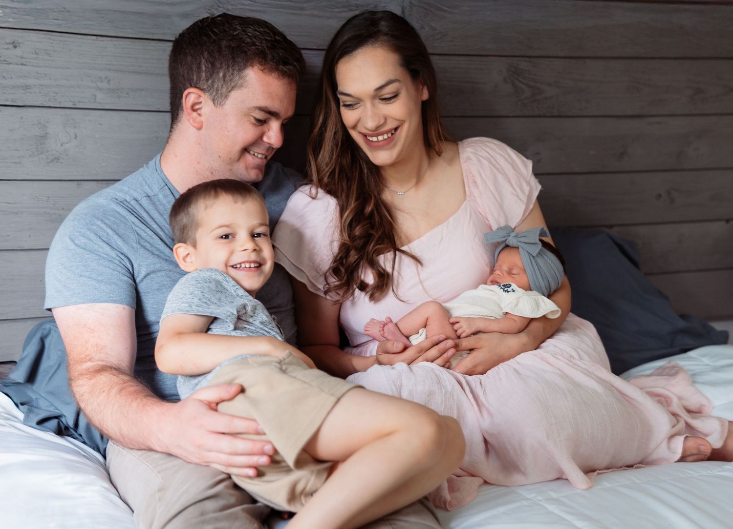 A family shares an intimate moment together on a bed with gray wood paneling in the background.