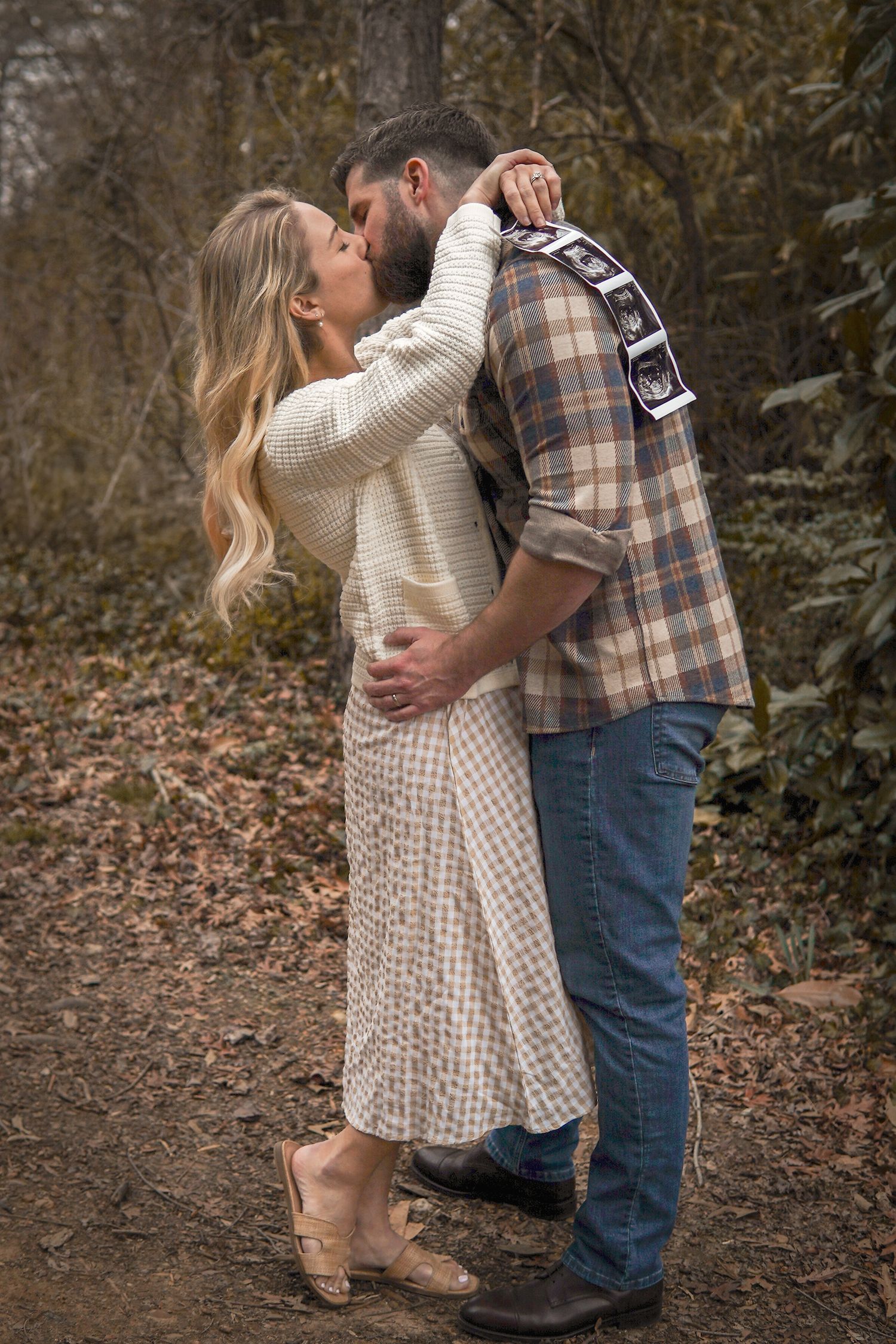A romantic couple embraces on a wooded autumn path, wearing cozy sweaters and casual attire.
