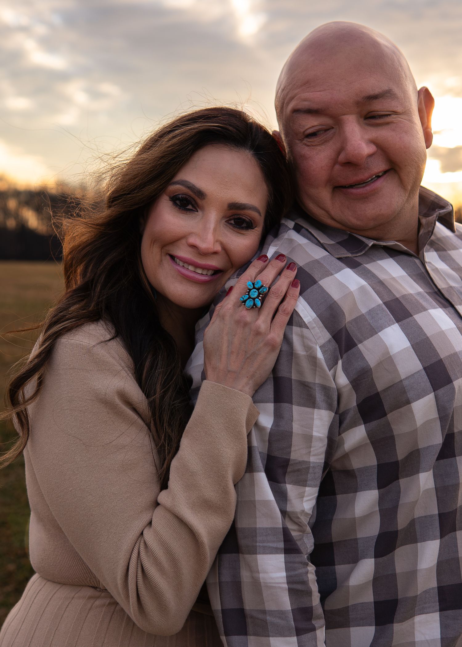 Smiling couple embracing at sunset in open field, woman wearing turquoise ring and beige sweater.
