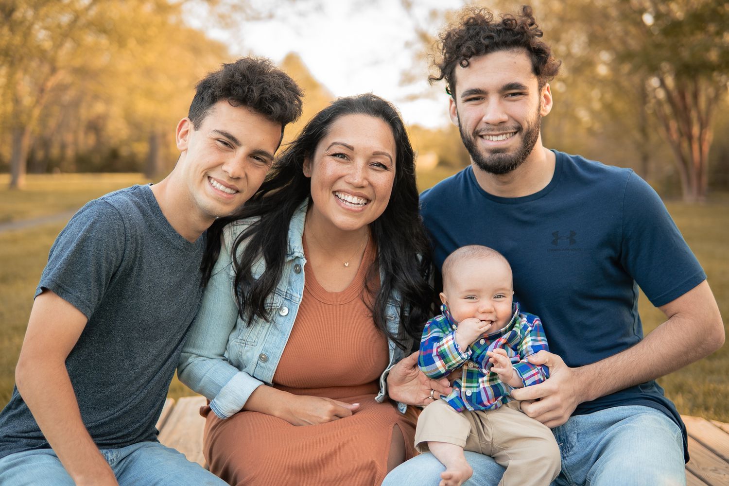 Family of four smiling outdoors in autumn park, including two teens, a woman, and a baby in plaid shirt.
