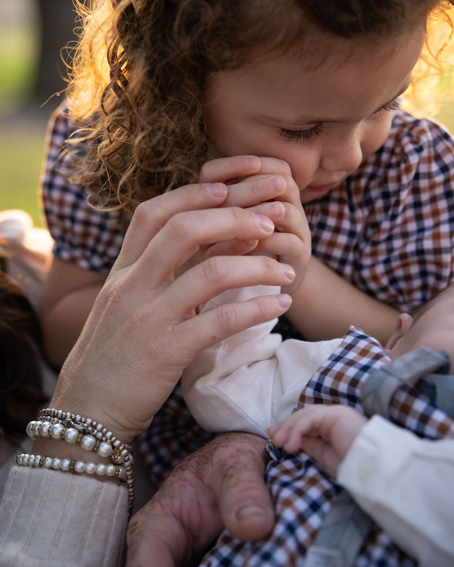 A curly-haired girl in gingham dress kisses an adult's hand while wearing pearl bracelet outdoors.