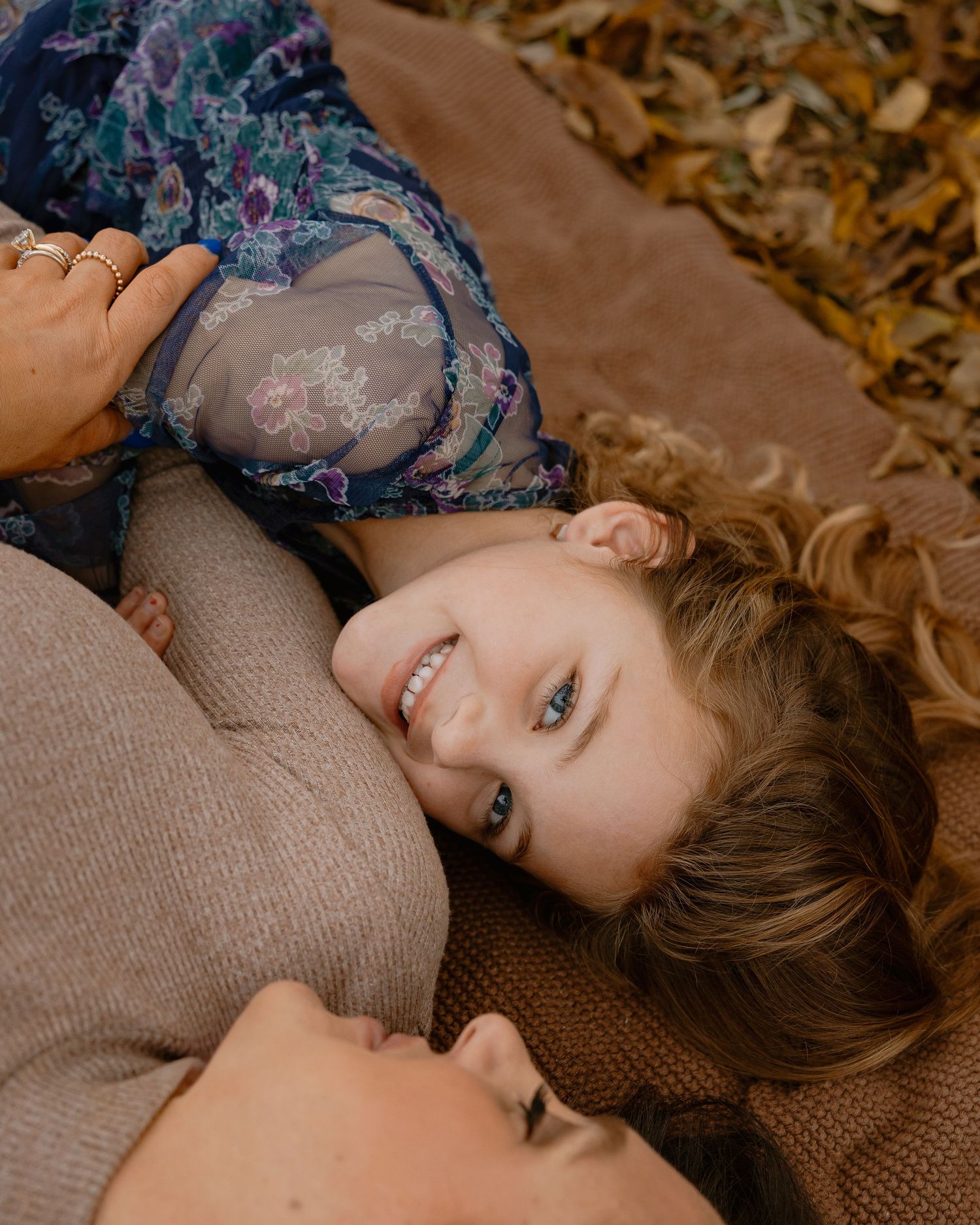A smiling child with blue eyes rests on a woman's lap outdoors among autumn leaves in a warm fall setting.