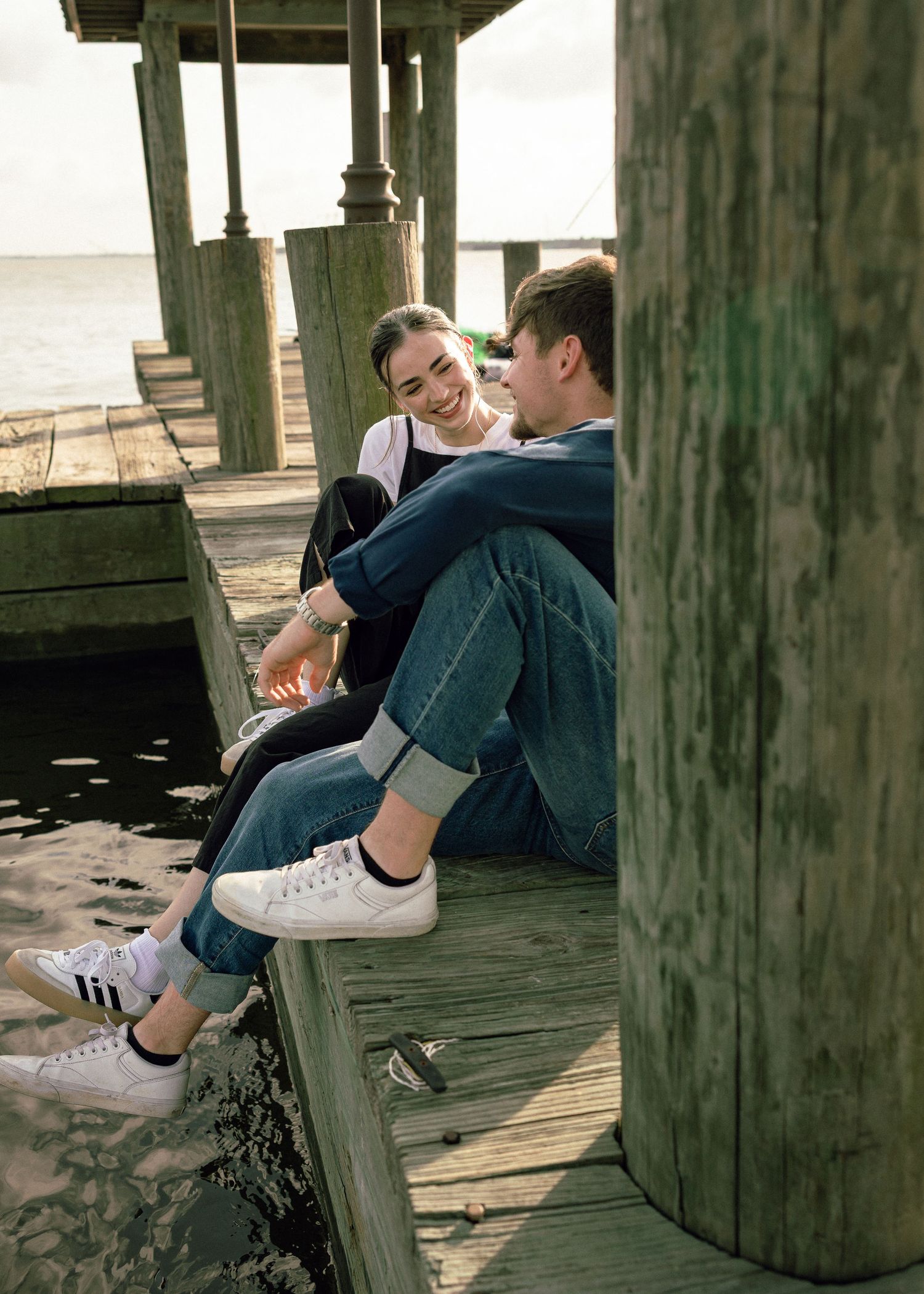 A young couple sits and smiles at each other on a wooden pier by the beach, wearing jeans and sneakers.