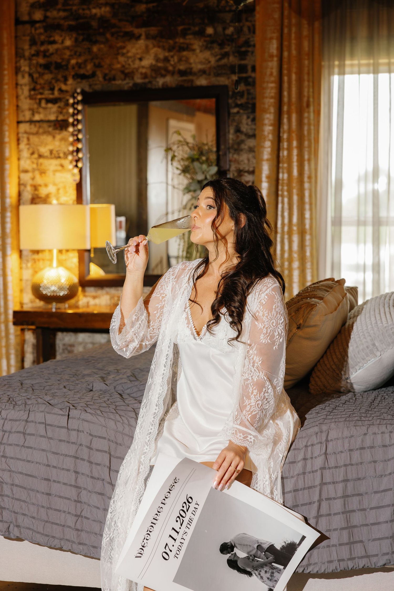 Bride in white lace robe sipping champagne while holding a wedding newspaper keepsake, seated on a hotel bed.