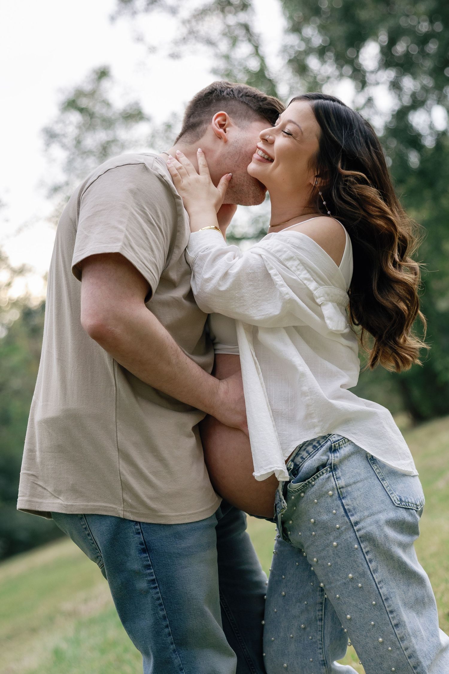 A happy couple shares a romantic moment outdoors, with the pregnant woman smiling as her partner leans in for a kiss.