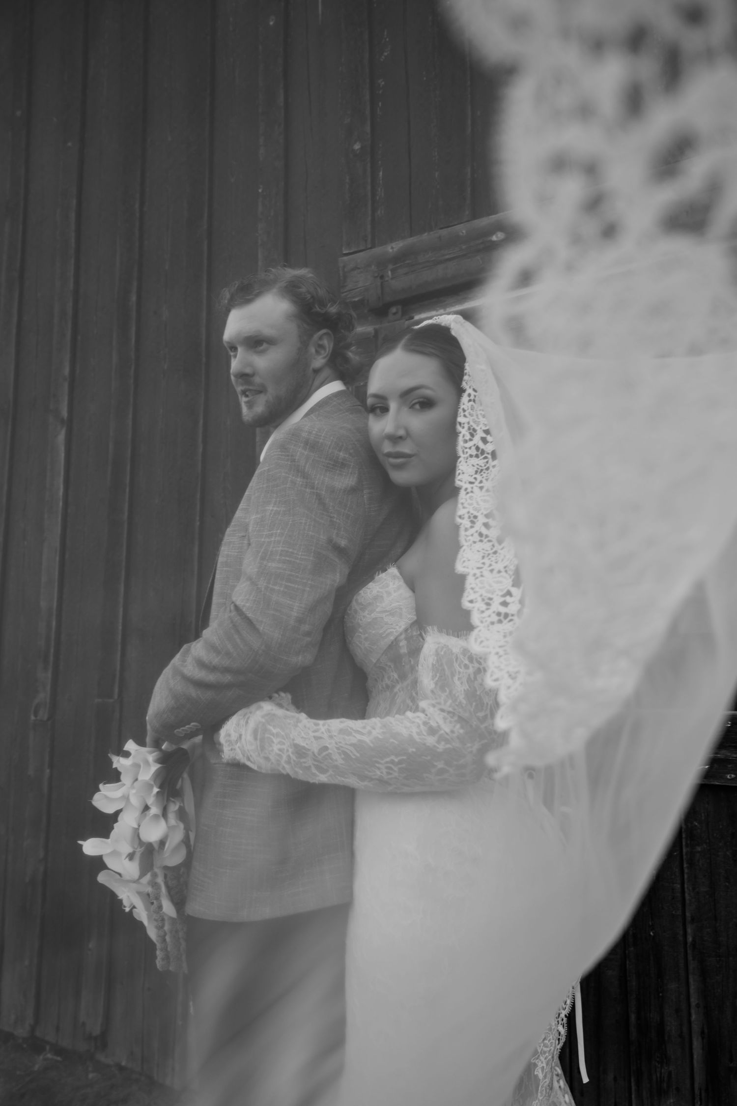 Black and white wedding photo of bride in lace veil embracing groom in suit against rustic wooden wall.