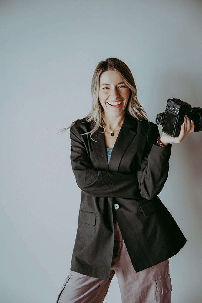 Smiling woman in black blazer holding a camera, posing confidently against a light gray background.