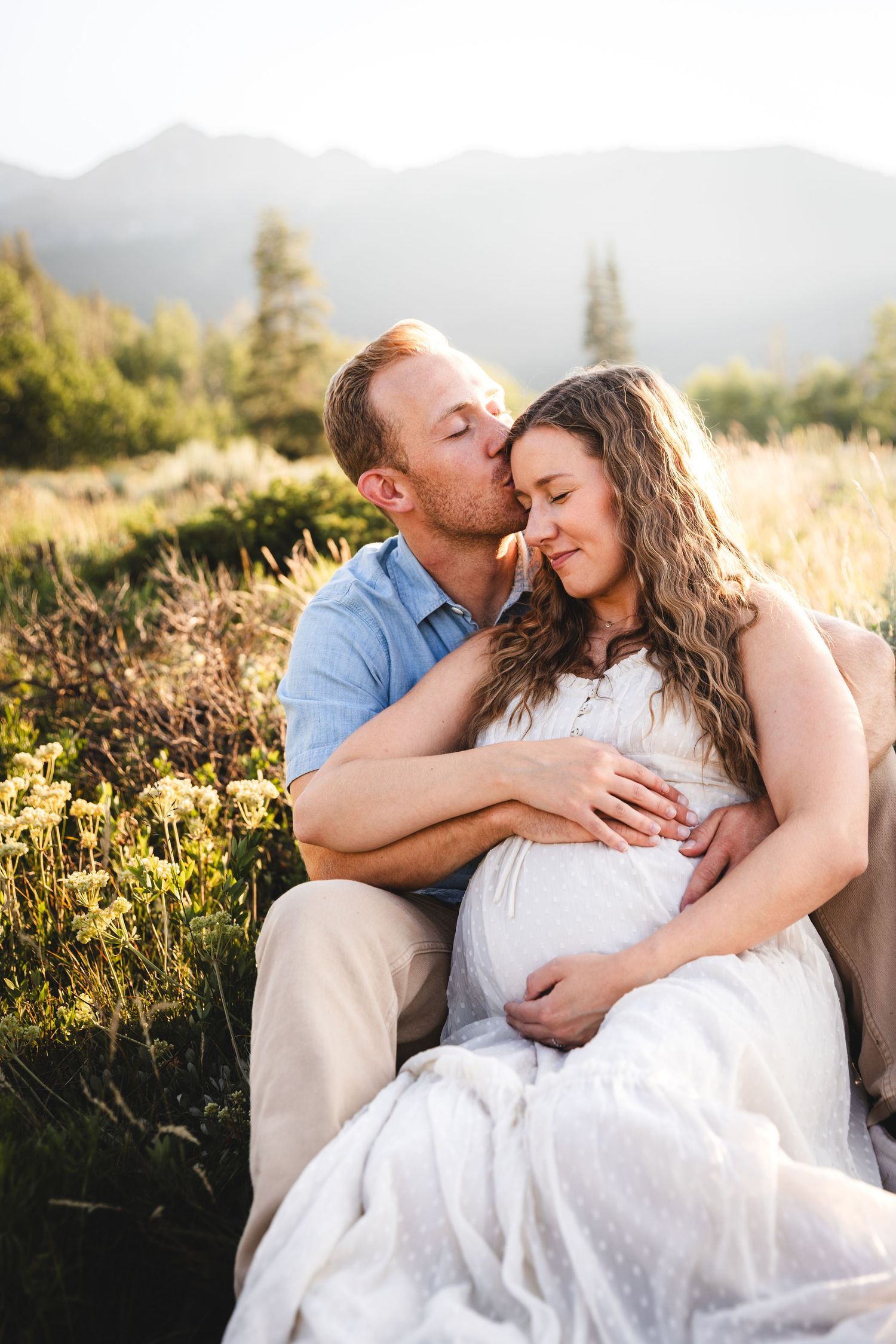 Couple sitting in mountain meadow, man kissing pregnant woman's temple while cradling her baby bump at golden hour. maternity