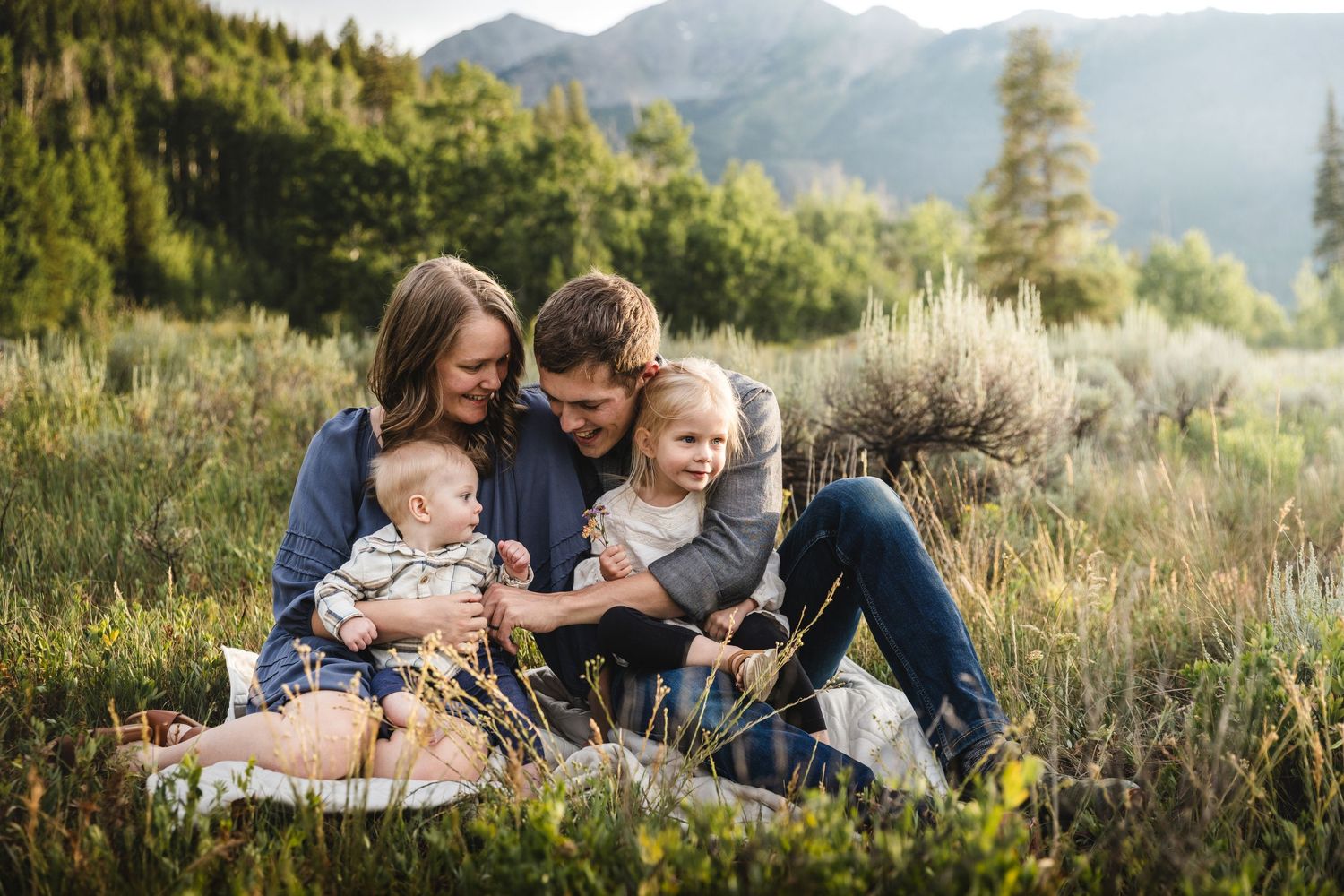 Family of four sitting in a meadow with mountain views, parents smiling at baby and toddler in golden hour light.