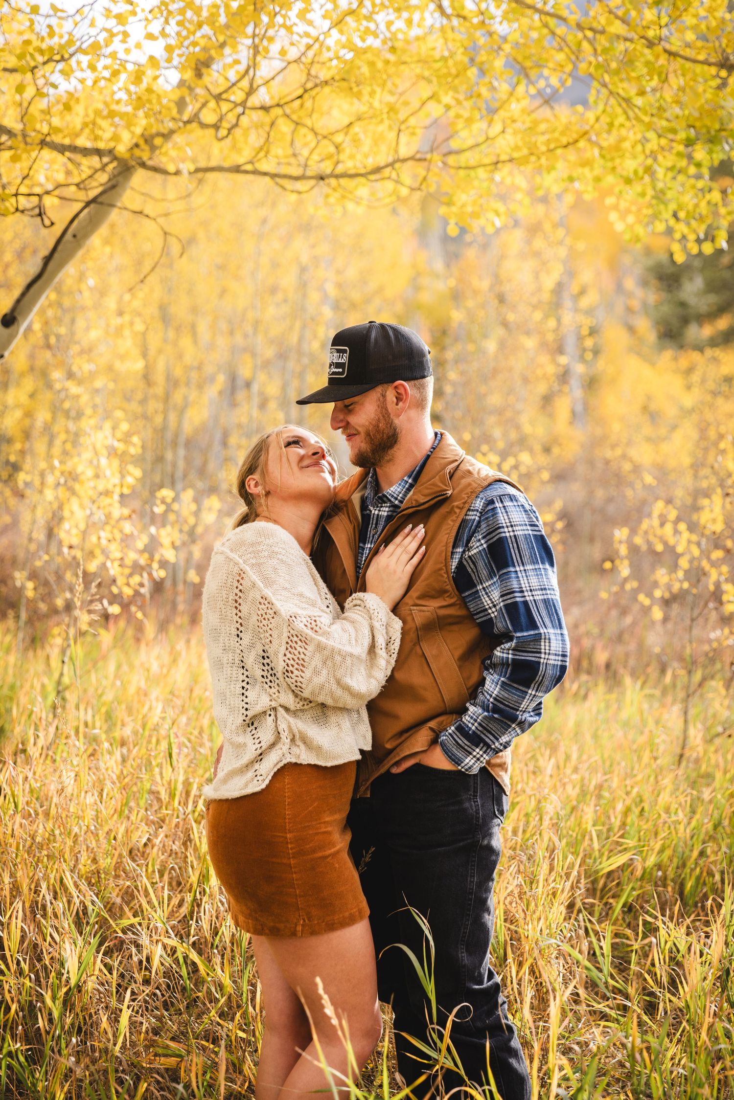 Couple embracing in golden autumn aspen forest, woman in cream sweater and suede skirt, man in plaid shirt and vest.