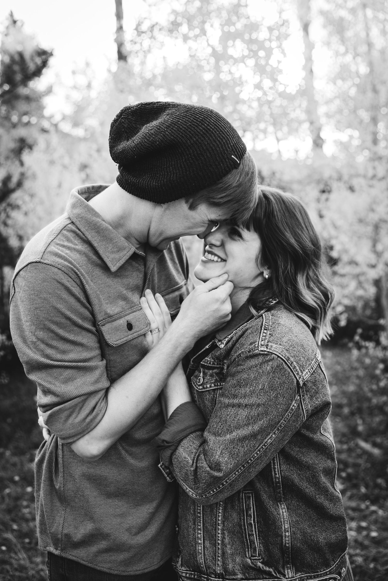 Black and white photo of a young couple sharing a romantic moment outdoors, man in beanie hat leaning in to kiss smiling woman.