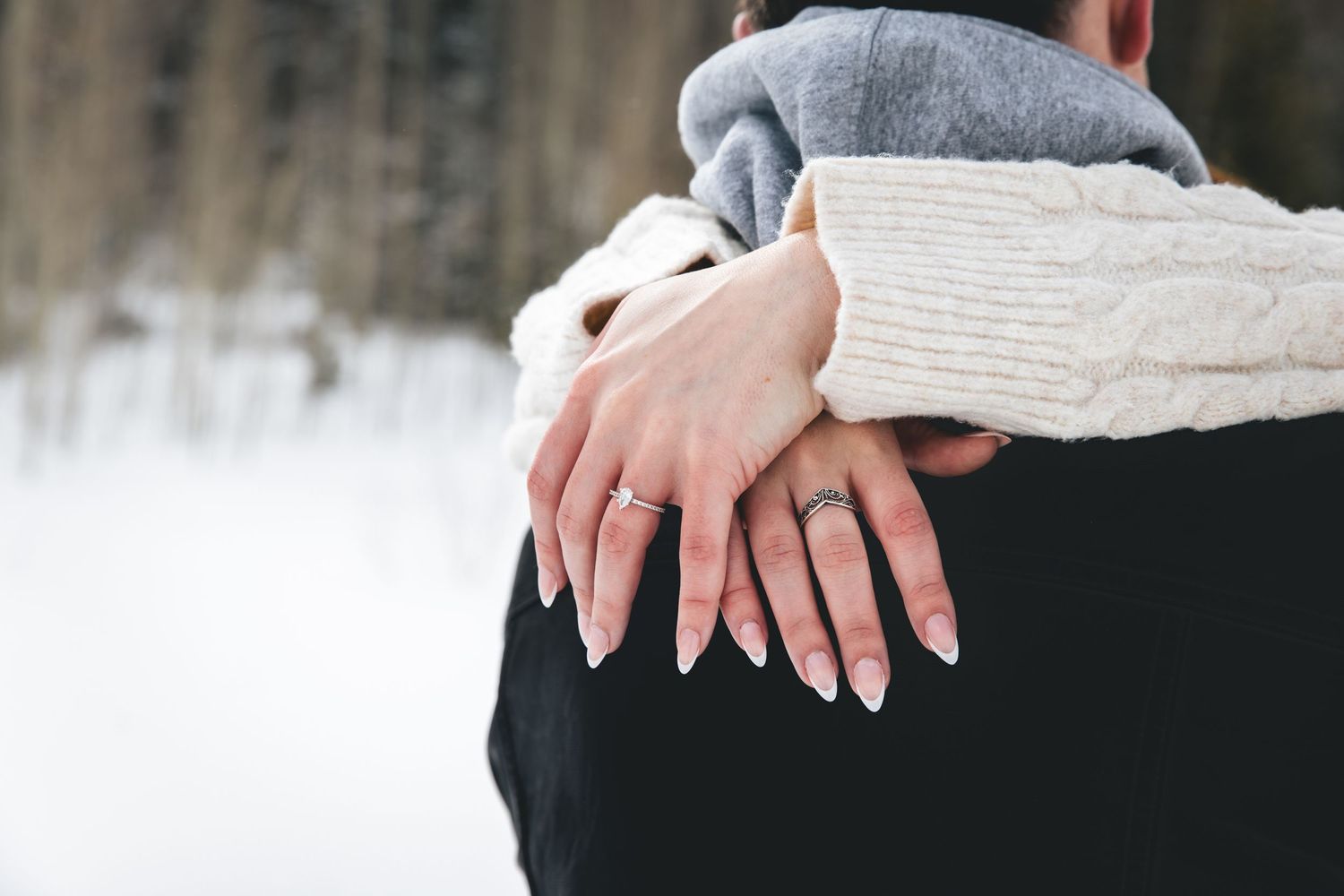 A close-up photo shows two hands with white manicured nails holding a small object between them. engagement ring
