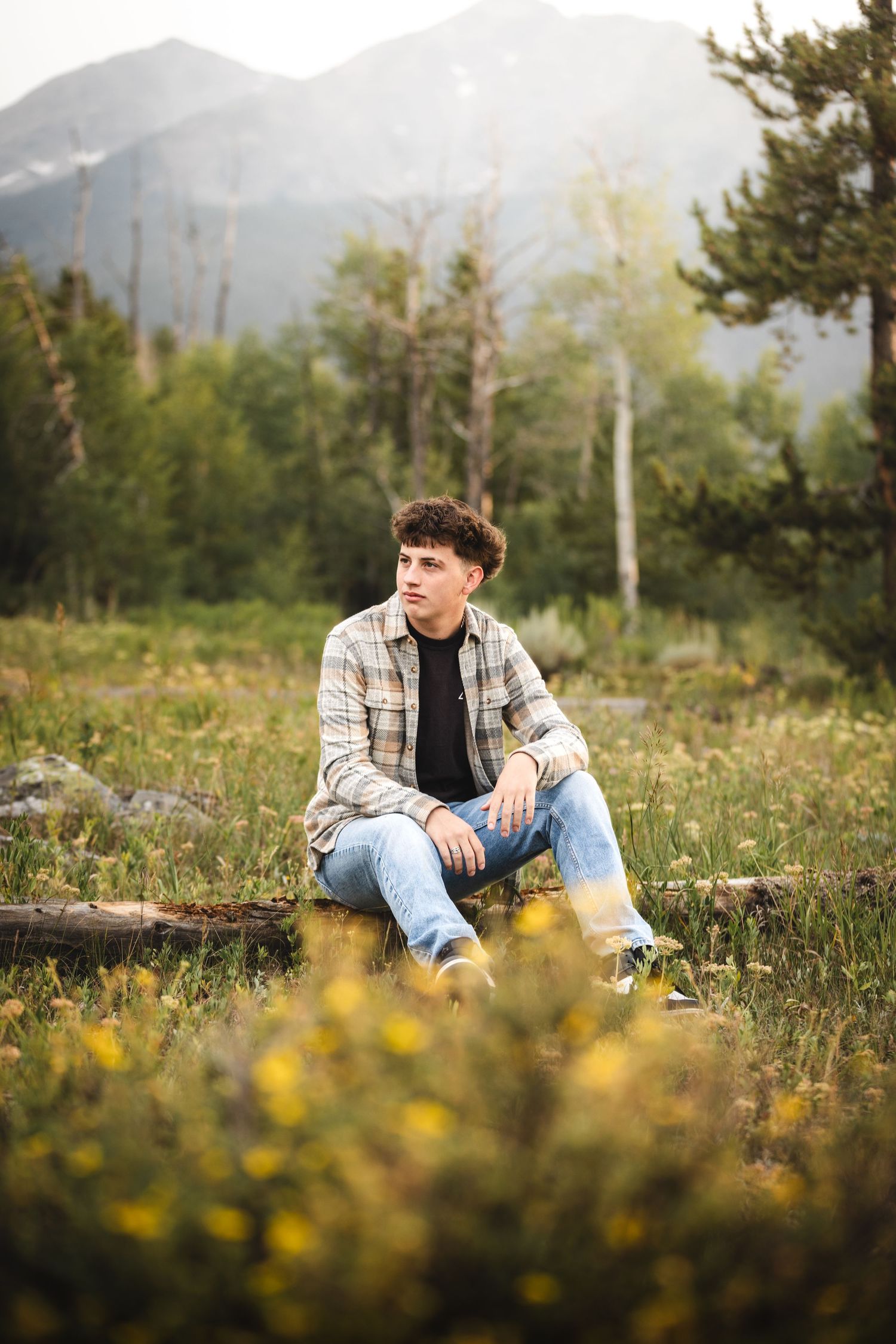 A person in plaid shirt and jeans sits in a meadow with yellow wildflowers and mountains in the background. - senior photos
