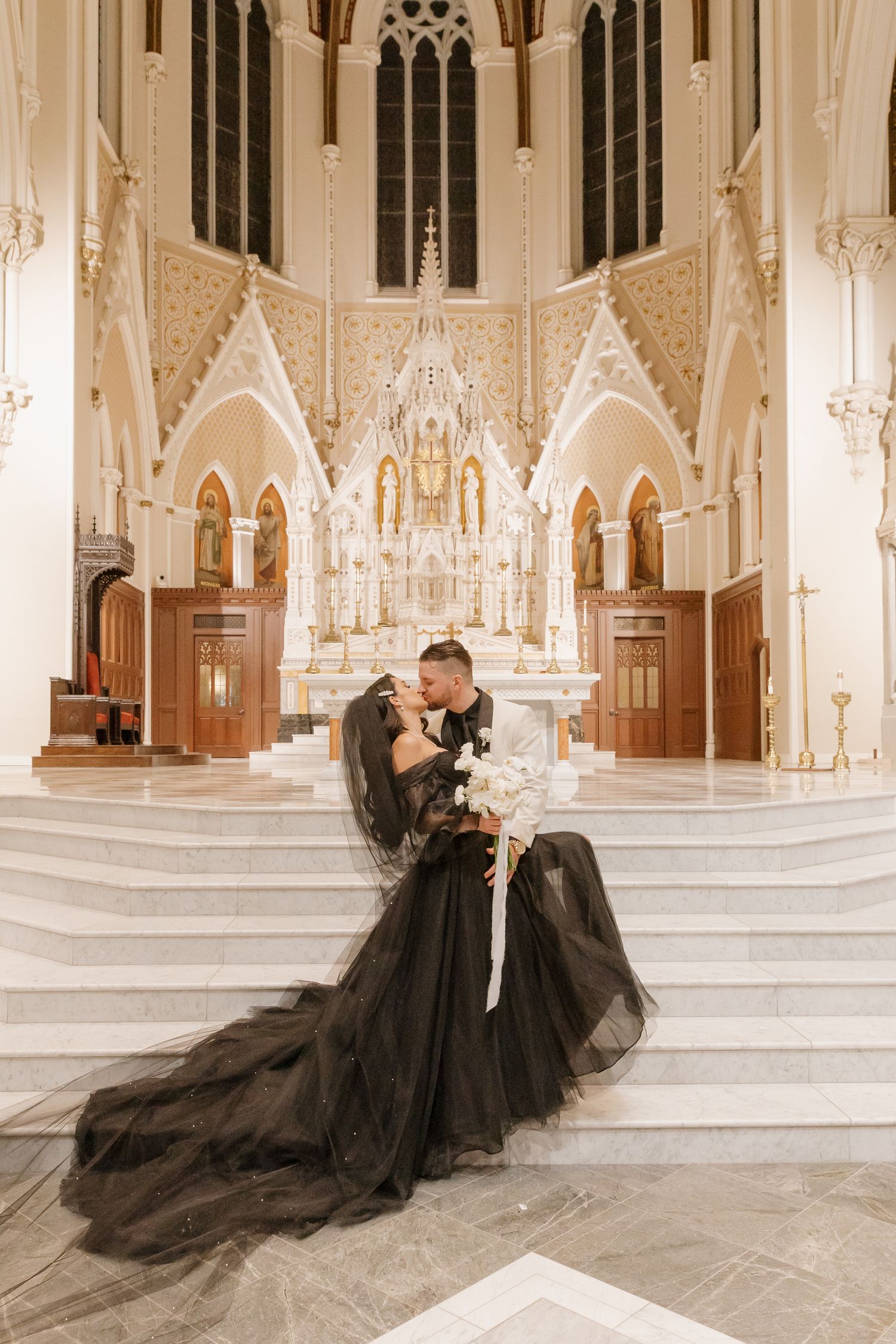 A couple in elegantly dressed in black embracing on white marble steps inside an ornate gothic cathedral with arched windows.