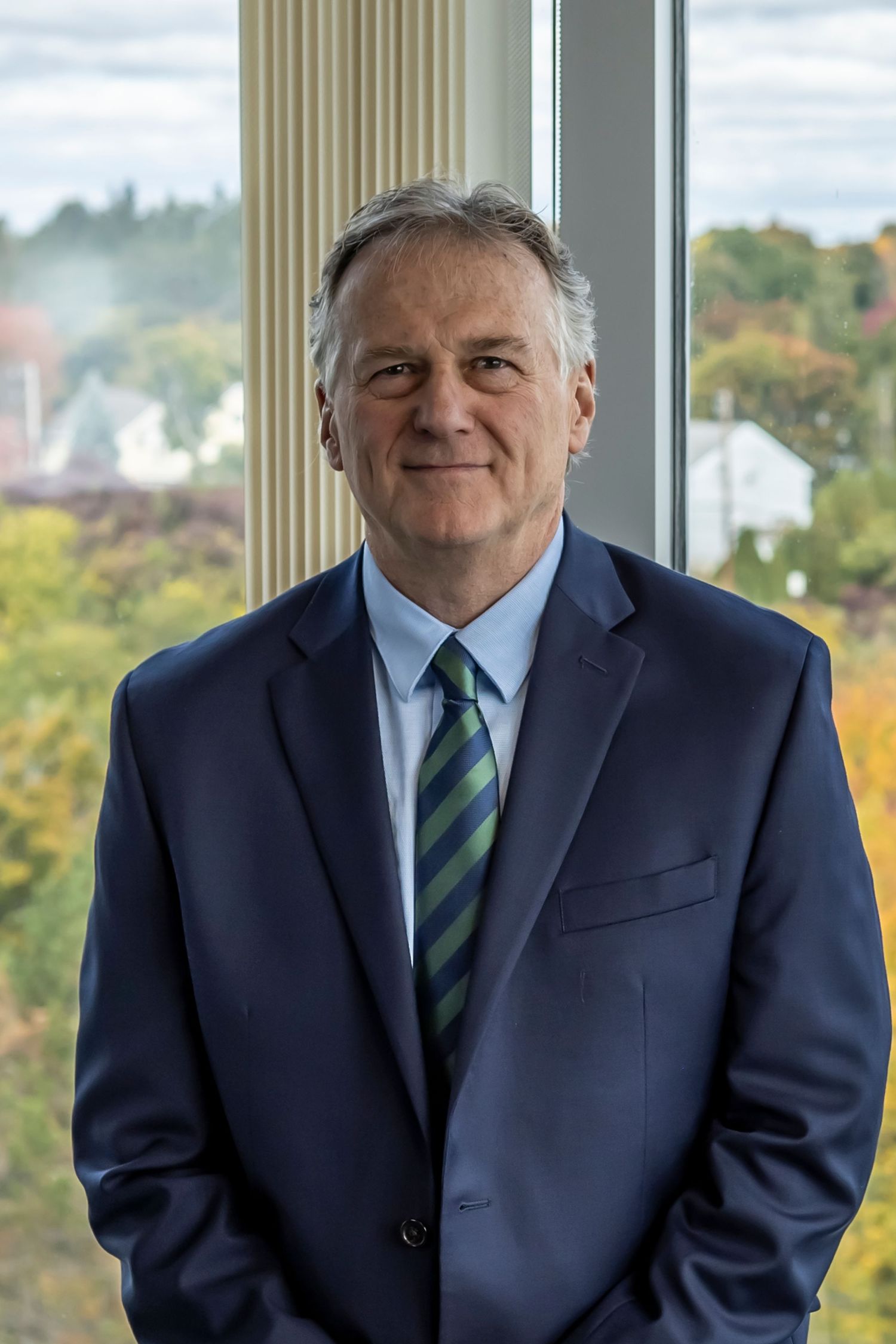 Professional headshot of someone in a navy suit and striped tie standing by a window with autumn foliage visible outside.