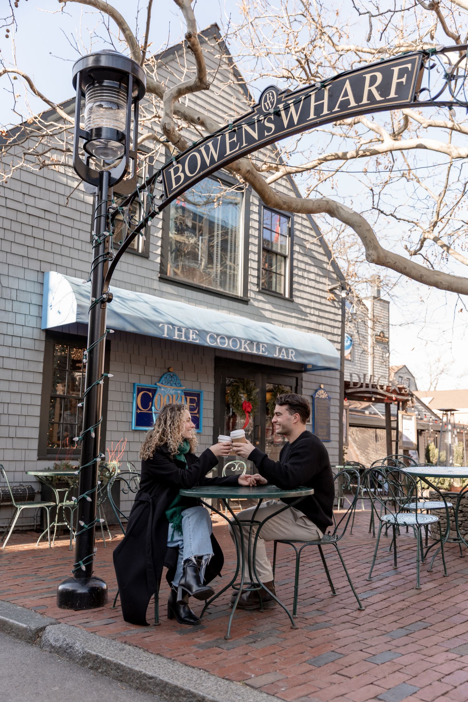 Diners enjoy outdoor seating at a cafe under the Bowens Wharf sign in Newport on a brick patio with vintage lamp post.