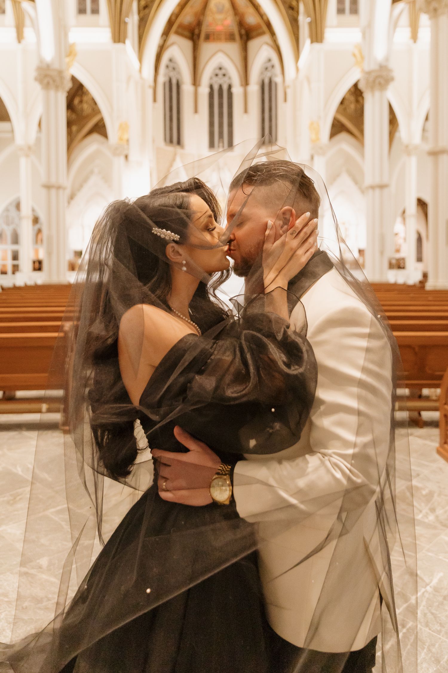A romantic wedding portrait inside a cathedral with a black gown bride and groom in white suit sharing a tender kiss.