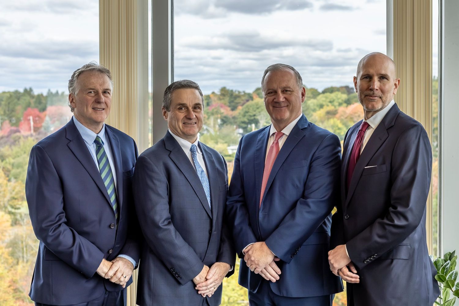 Four business professionals in suits stand together near a window overlooking colorful autumn trees.