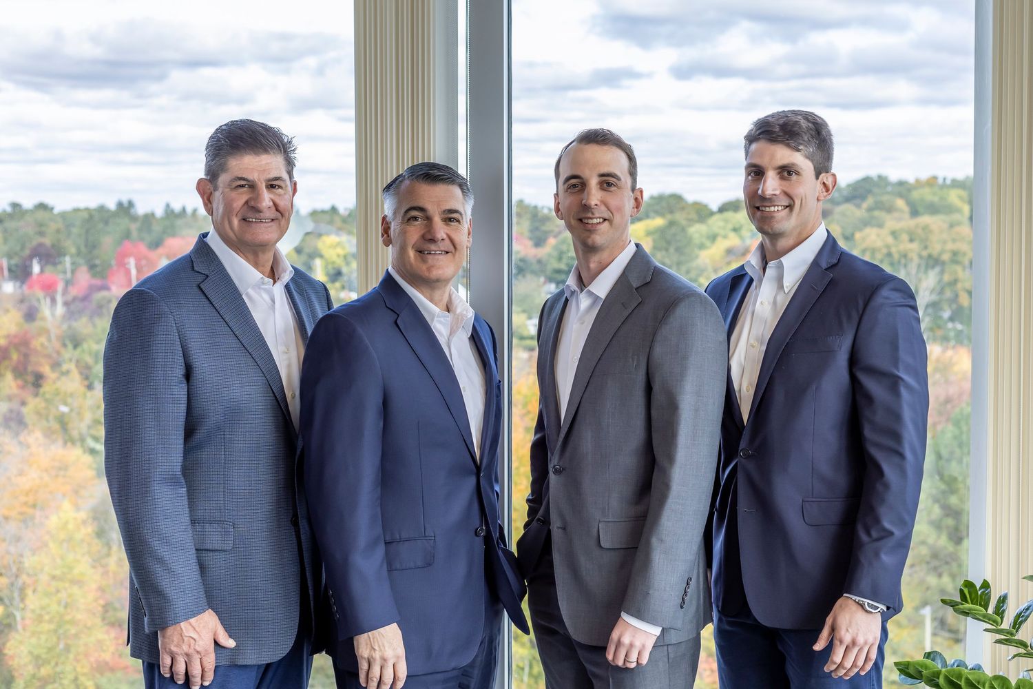 Four people in navy blue business suits stand together by a window with autumn foliage visible in the background.