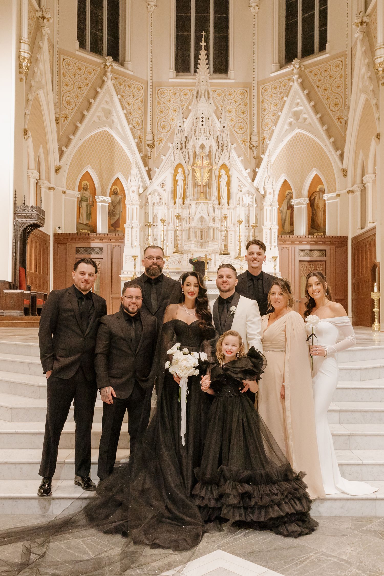Wedding party poses in elegant gothic cathedral with ornate white altar and arches wearing black and white formal attire.