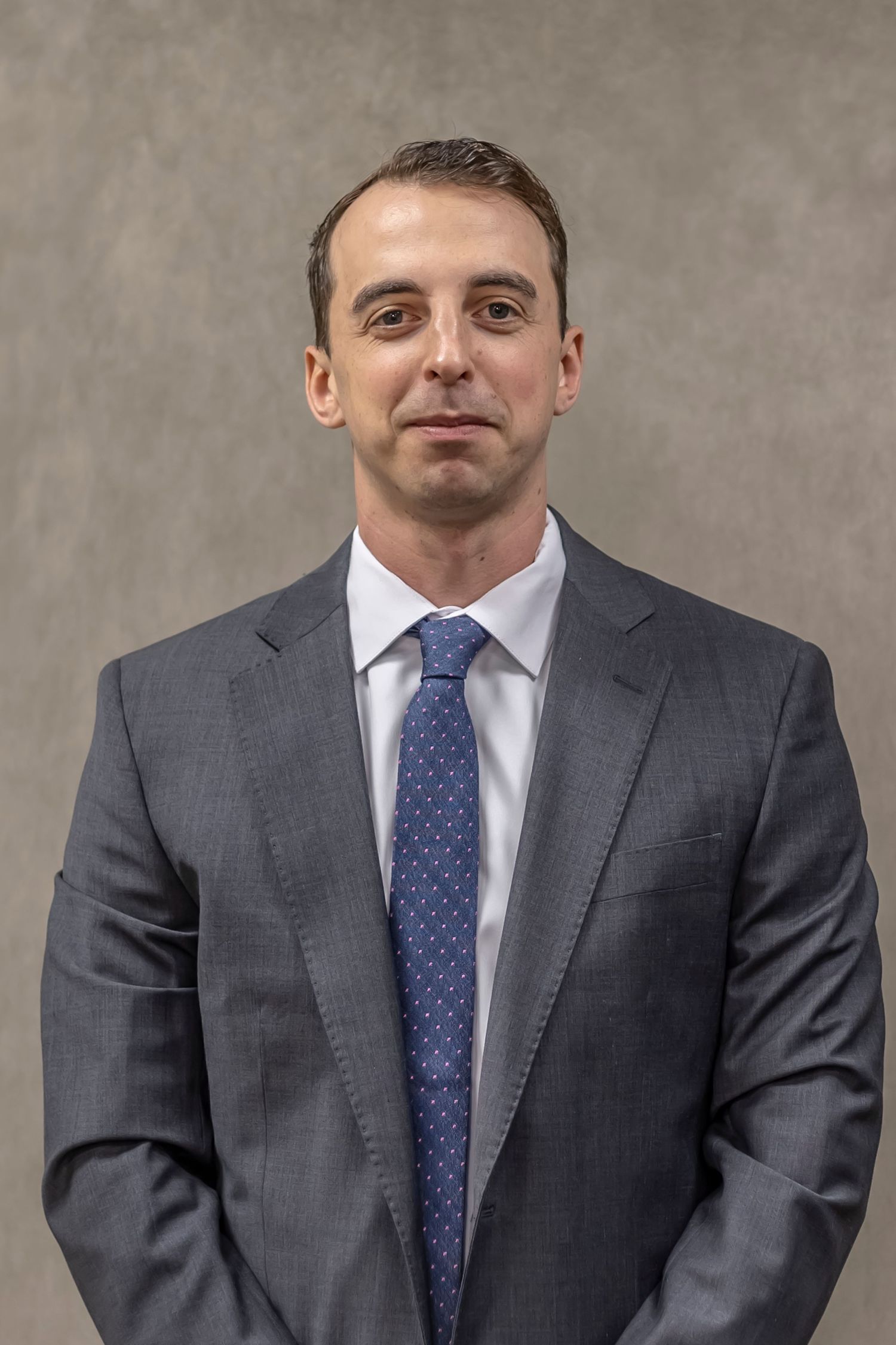 Professional headshot portrait of someone in a gray suit and blue polka dot tie against a neutral backdrop.
