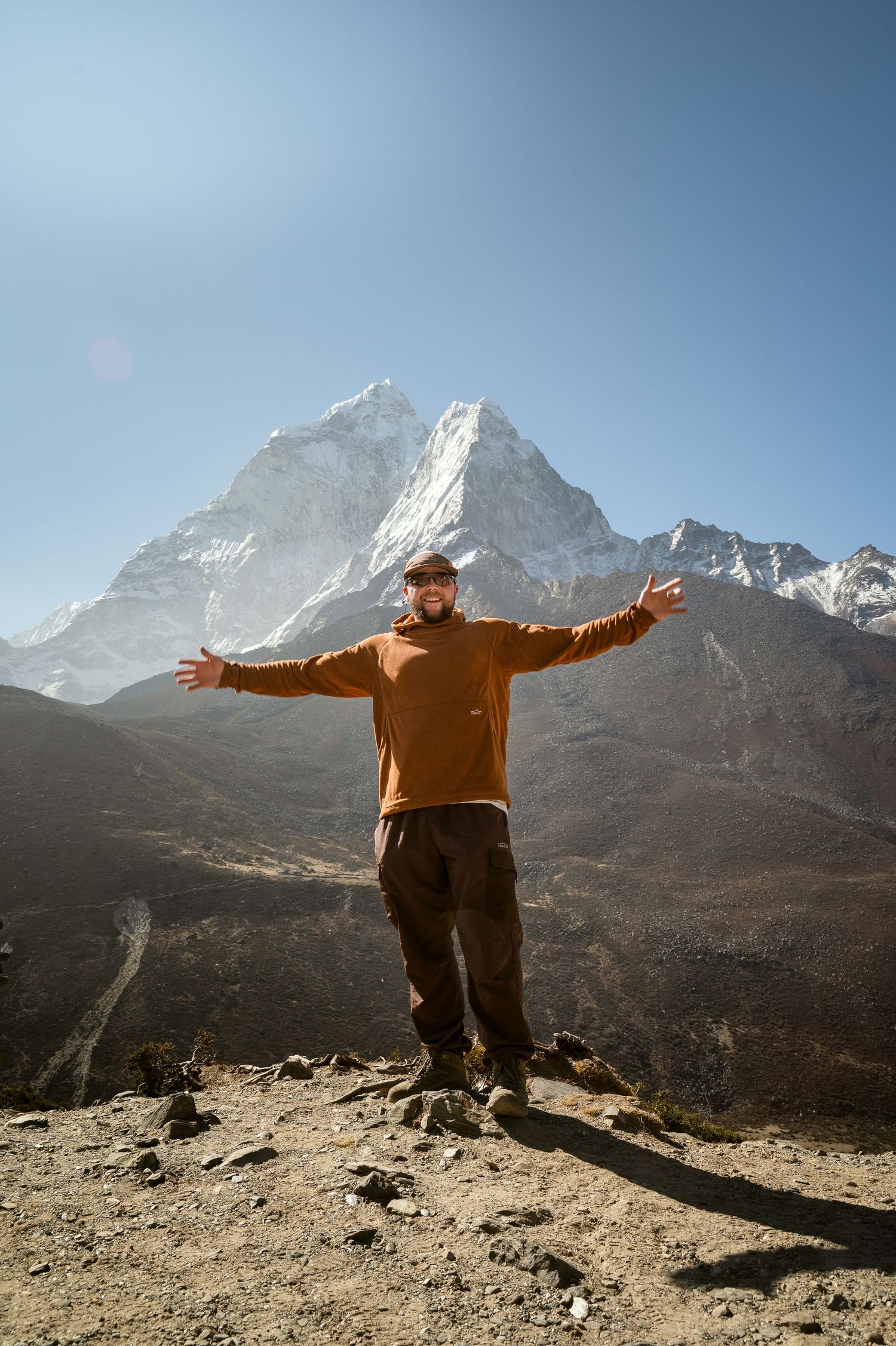Man in orange sweater stands with arms outstretched on rocky terrain with snow-capped Himalayan mountain peak behind him.
