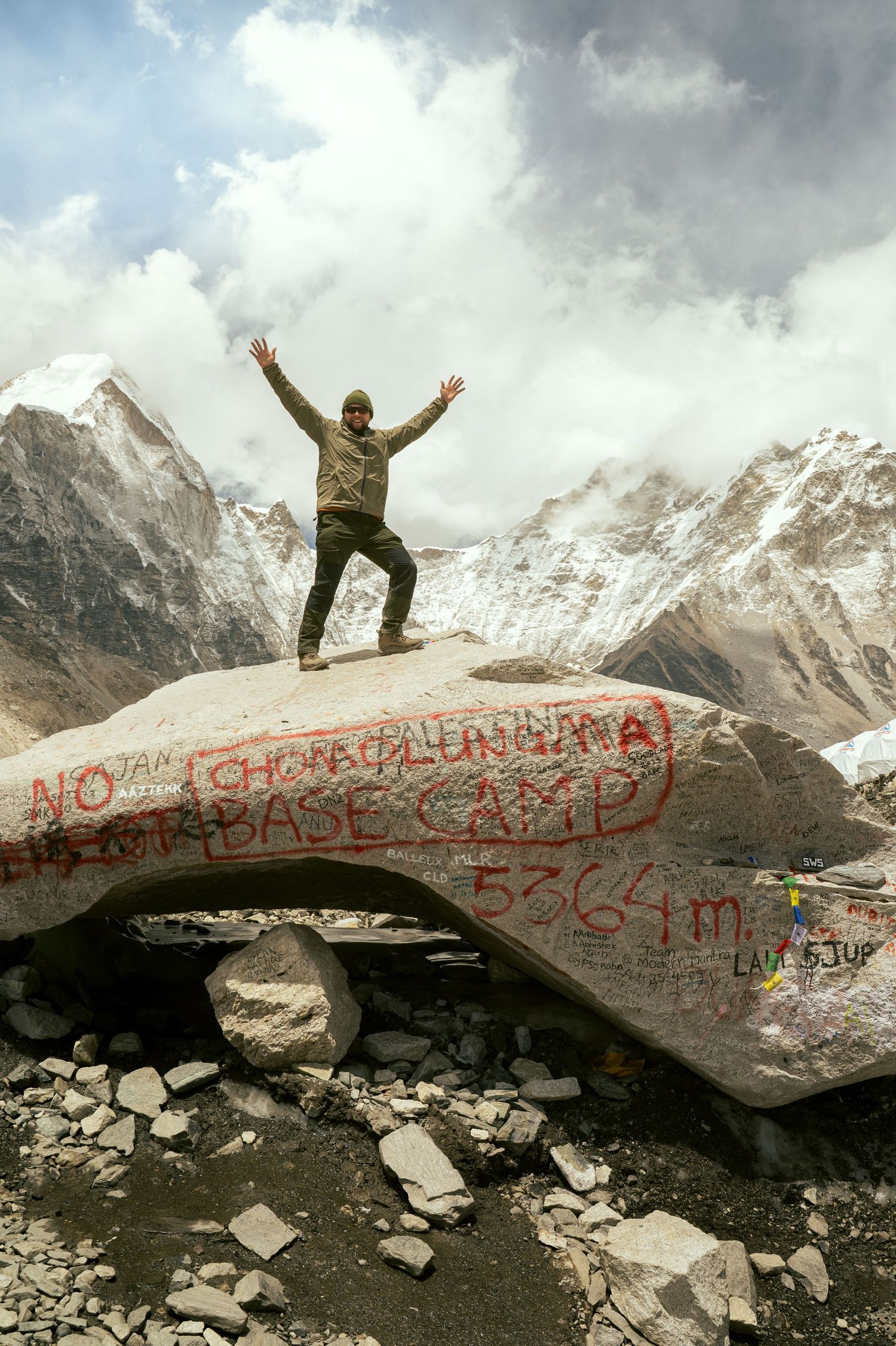 A person stands triumphantly atop a graffiti-covered rock formation with snowy Himalayan peaks in the background.