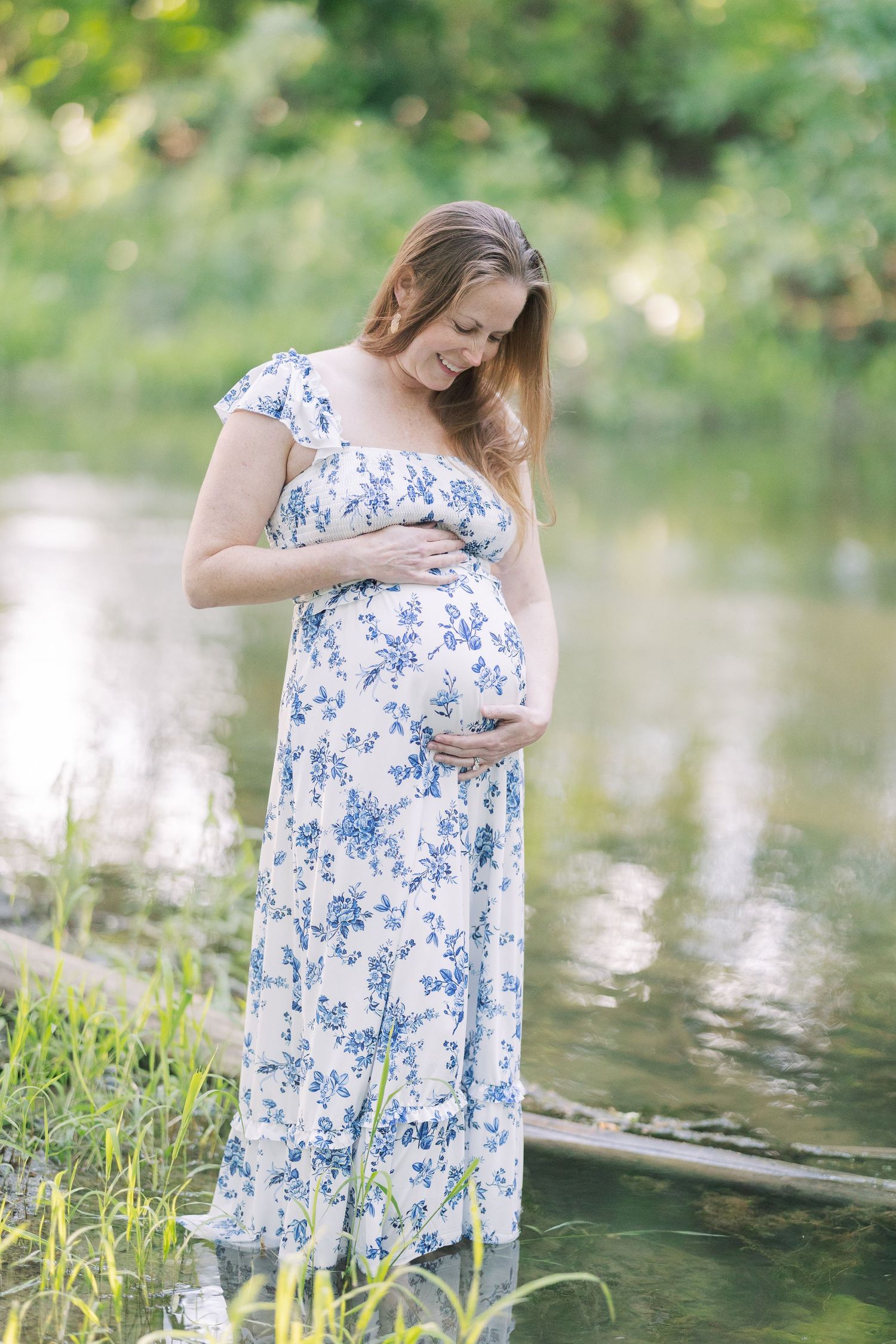 Pregnant woman in blue floral maxi dress cradling her baby bump outdoors near a serene pond surrounded by lush greenery.