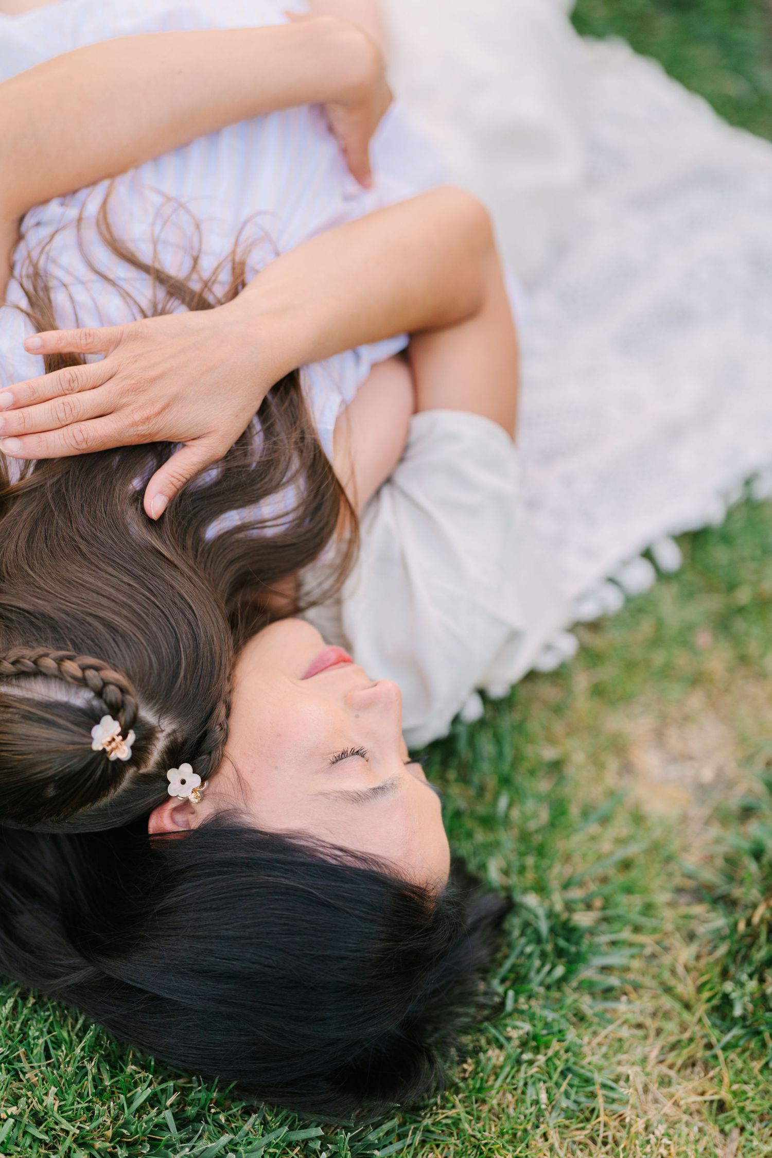Young woman with dark wavy hair and braids lying on green grass in a white dress, eyes closed peacefully.