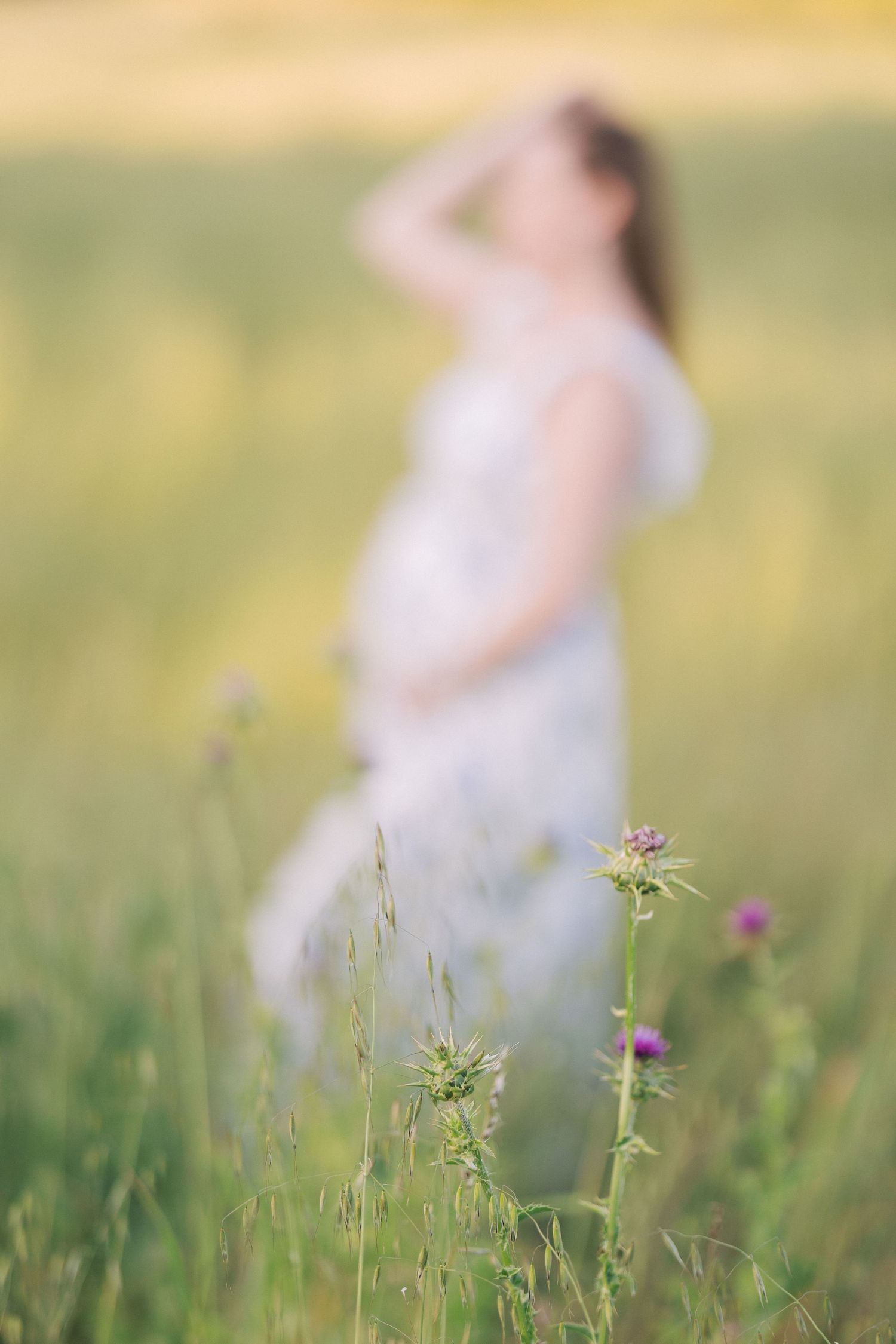 Pregnant woman in white dress standing in a sunny meadow with purple wildflowers in the foreground.