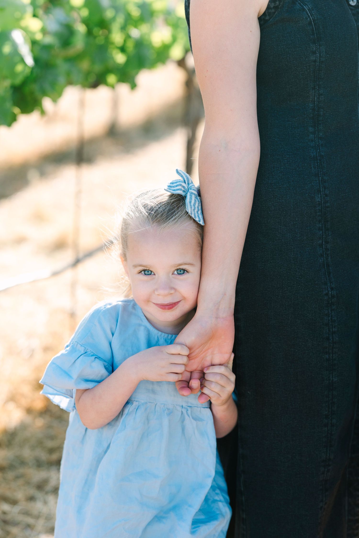 Small child in light blue dress holds adult's hand against natural backdrop.