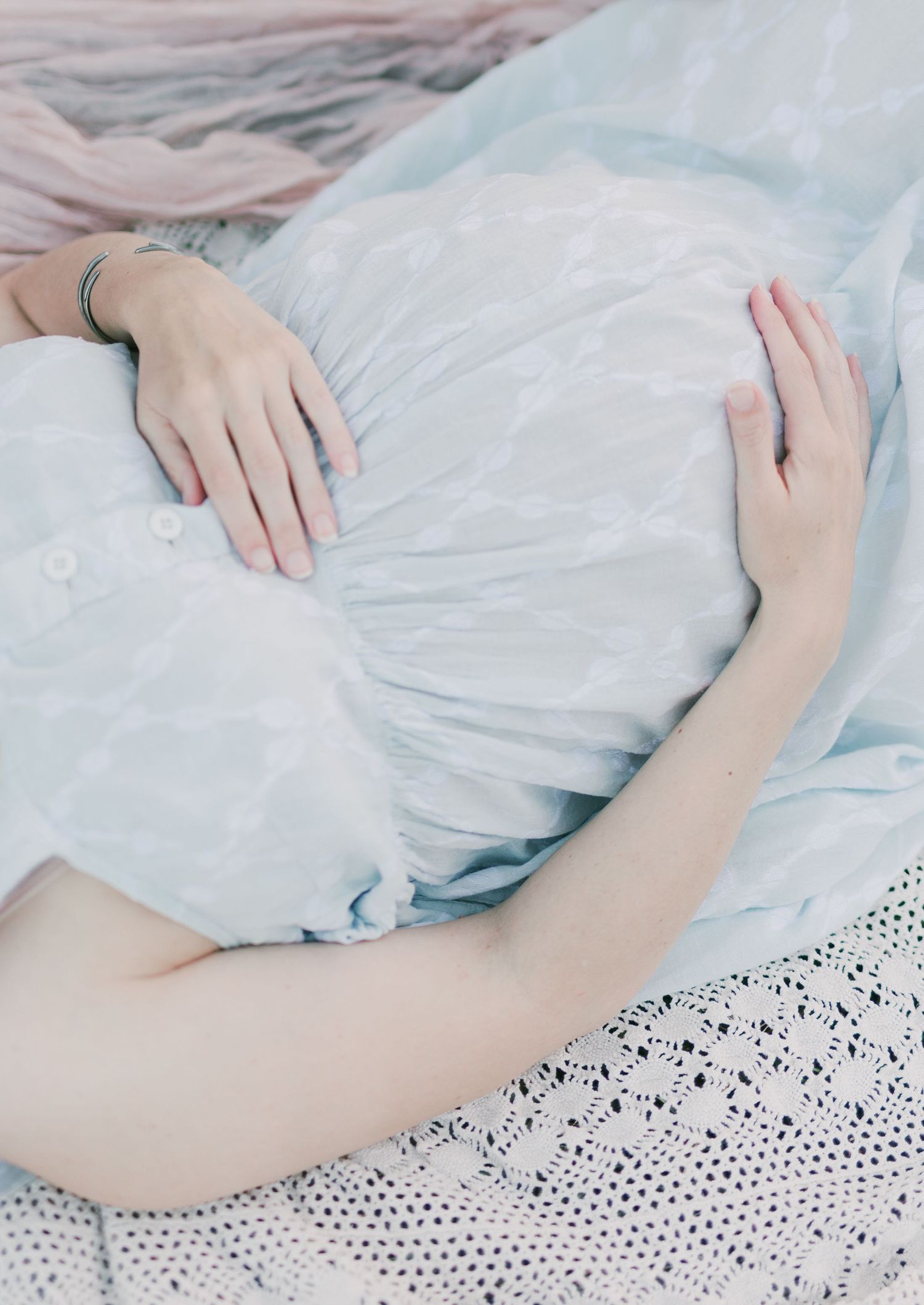 Pregnant woman in a light blue dress gently cradling her baby bump with both hands while lying down.