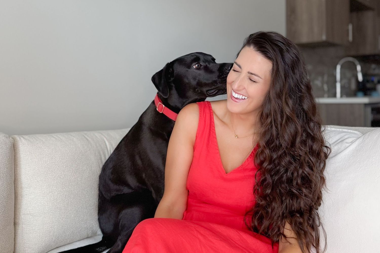 Candid photo of pet photographer and her black dog on a couch. Black dog is kissing her cheek and she is smiling with eyes closed