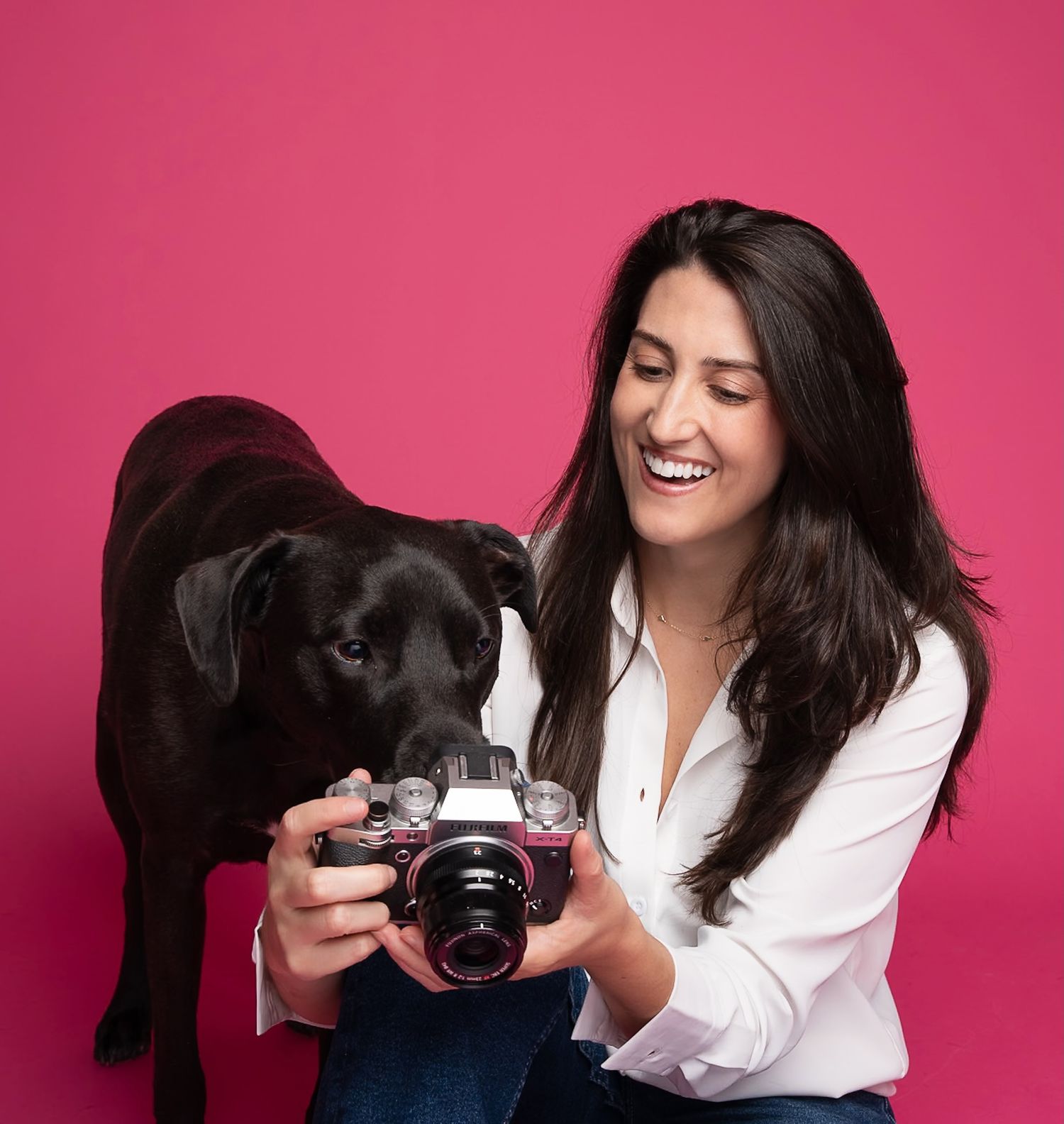 Pet photographer posing with a black dog and camera against a pink studio backdrop