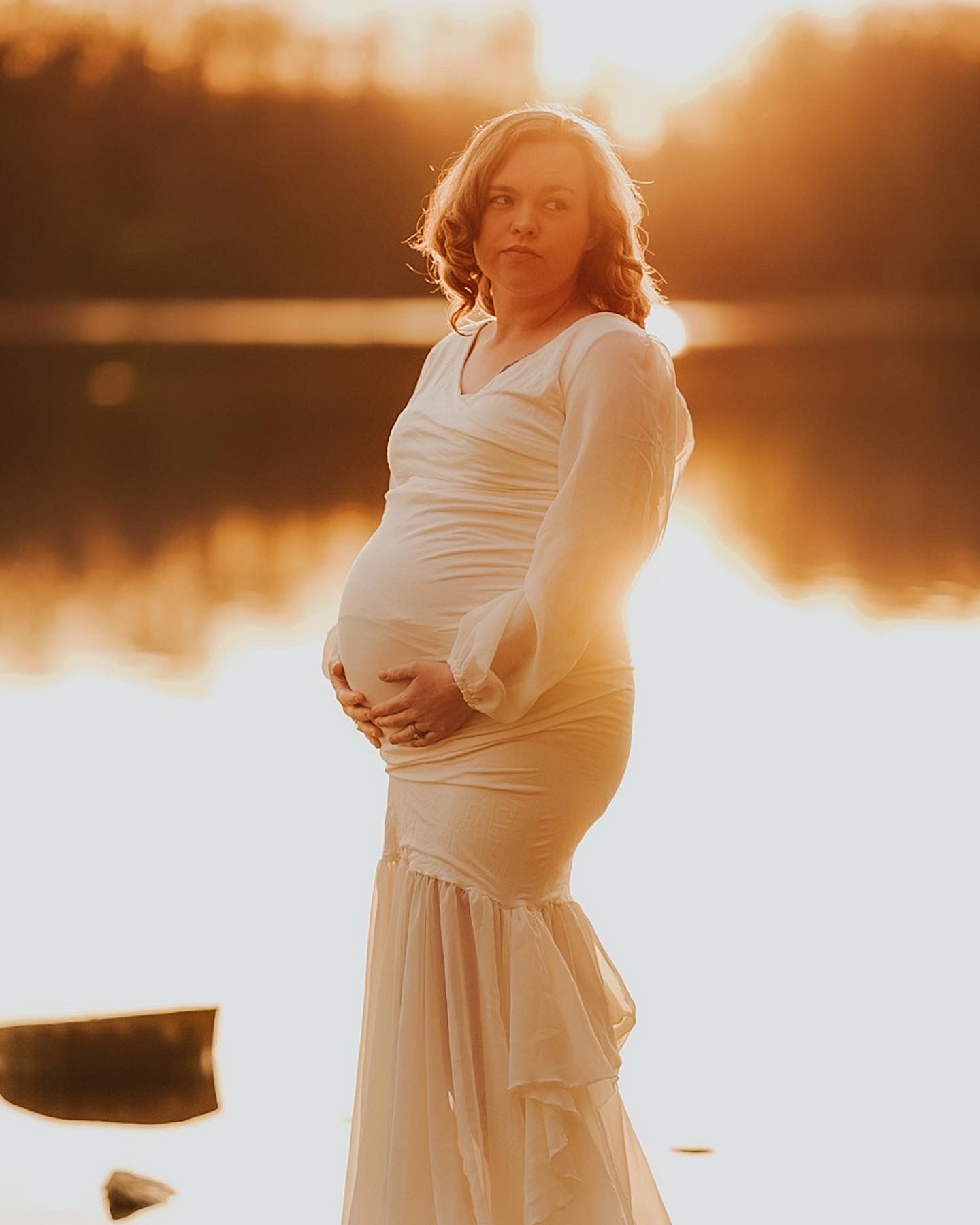 Maternity silhouettes at sunset by lake showing woman in flowing white dress.