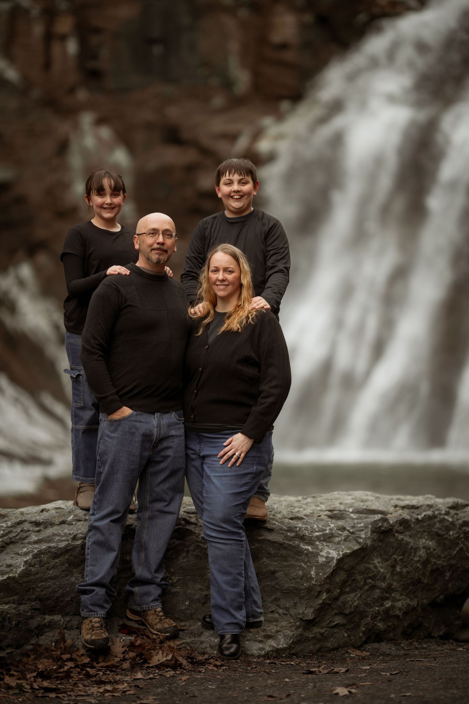 Family of four posing in front of a waterfall, wearing matching black sweaters and blue jeans outdoors.