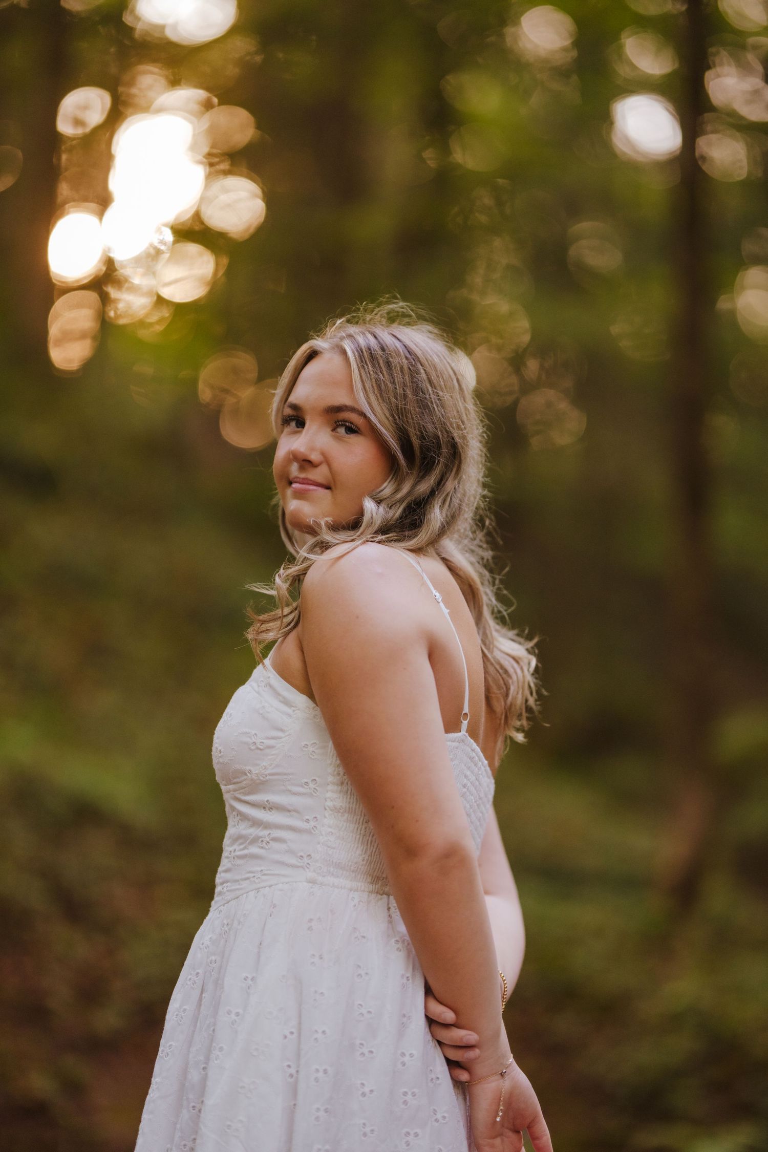 Woman in white sundress poses against blurred forest backdrop with bokeh lighting.