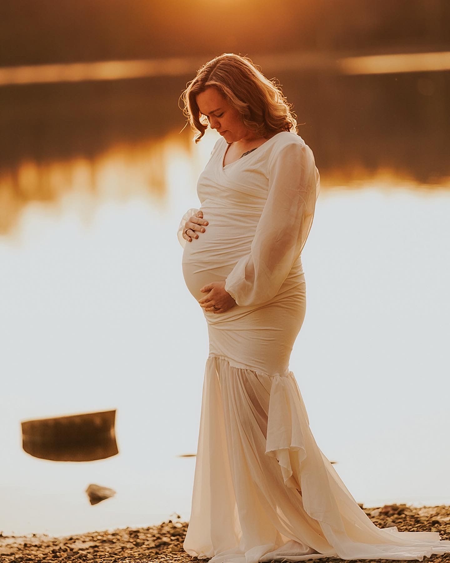 A serene maternity photoshoot at sunset by a lake featuring a flowing white dress.