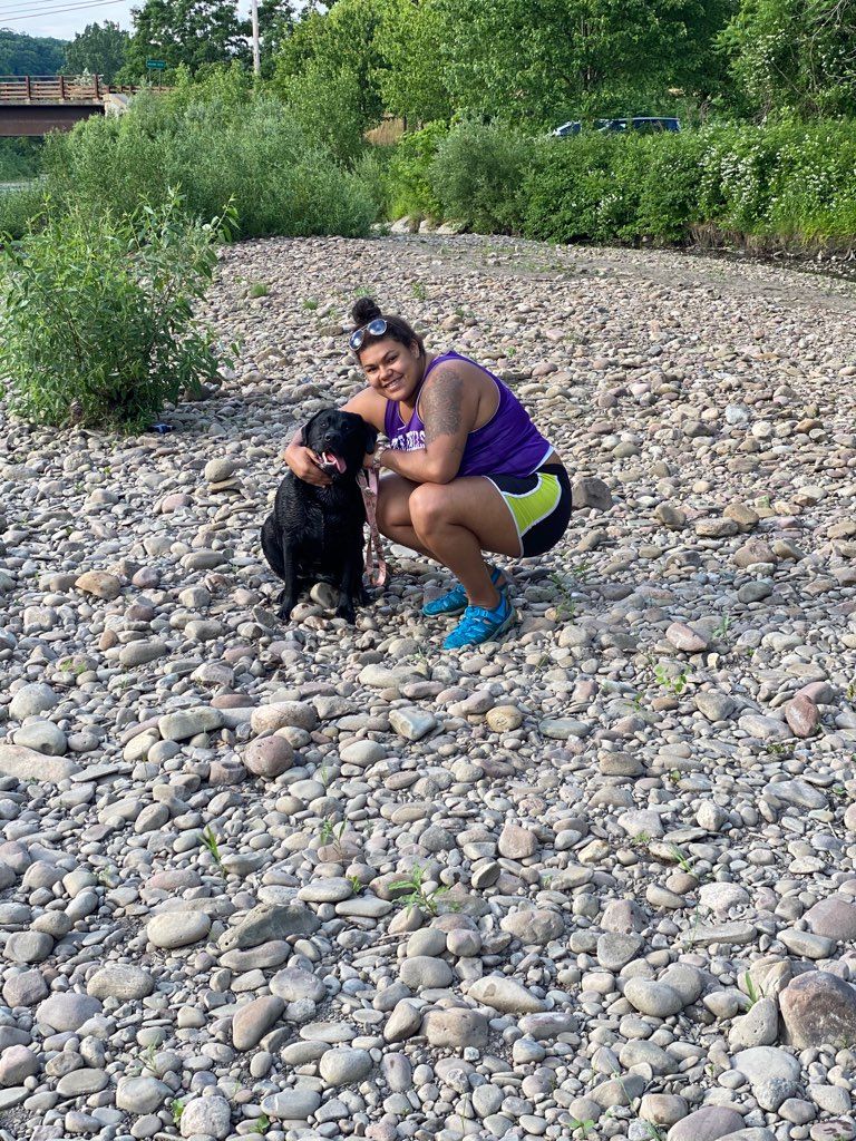 Someone in athletic wear crouching next to black dog on rocky riverbank.