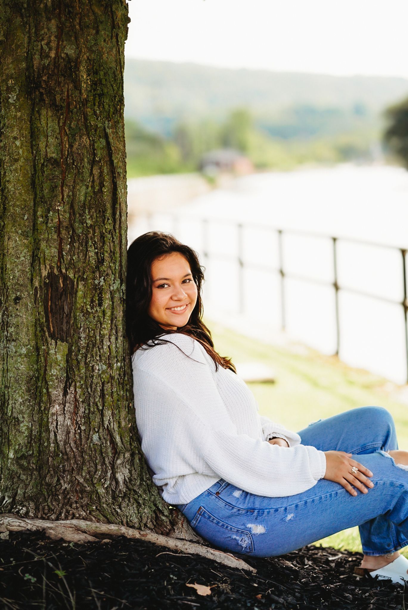 Someone relaxes against a tree trunk wearing jeans and a white sweater with a scenic backdrop.